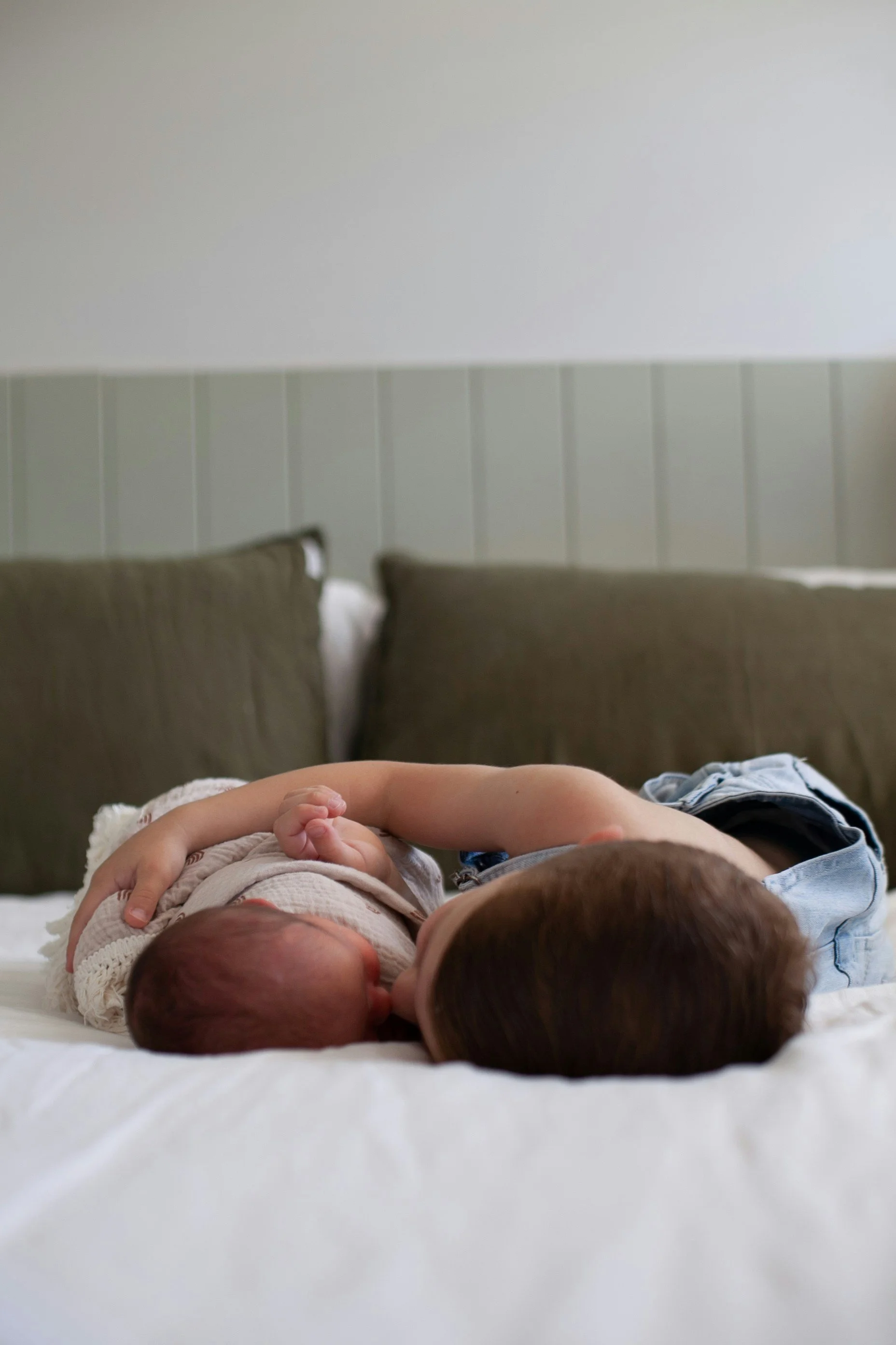 A newborn baby and a young child lying on a bed, with pillows against a wall.