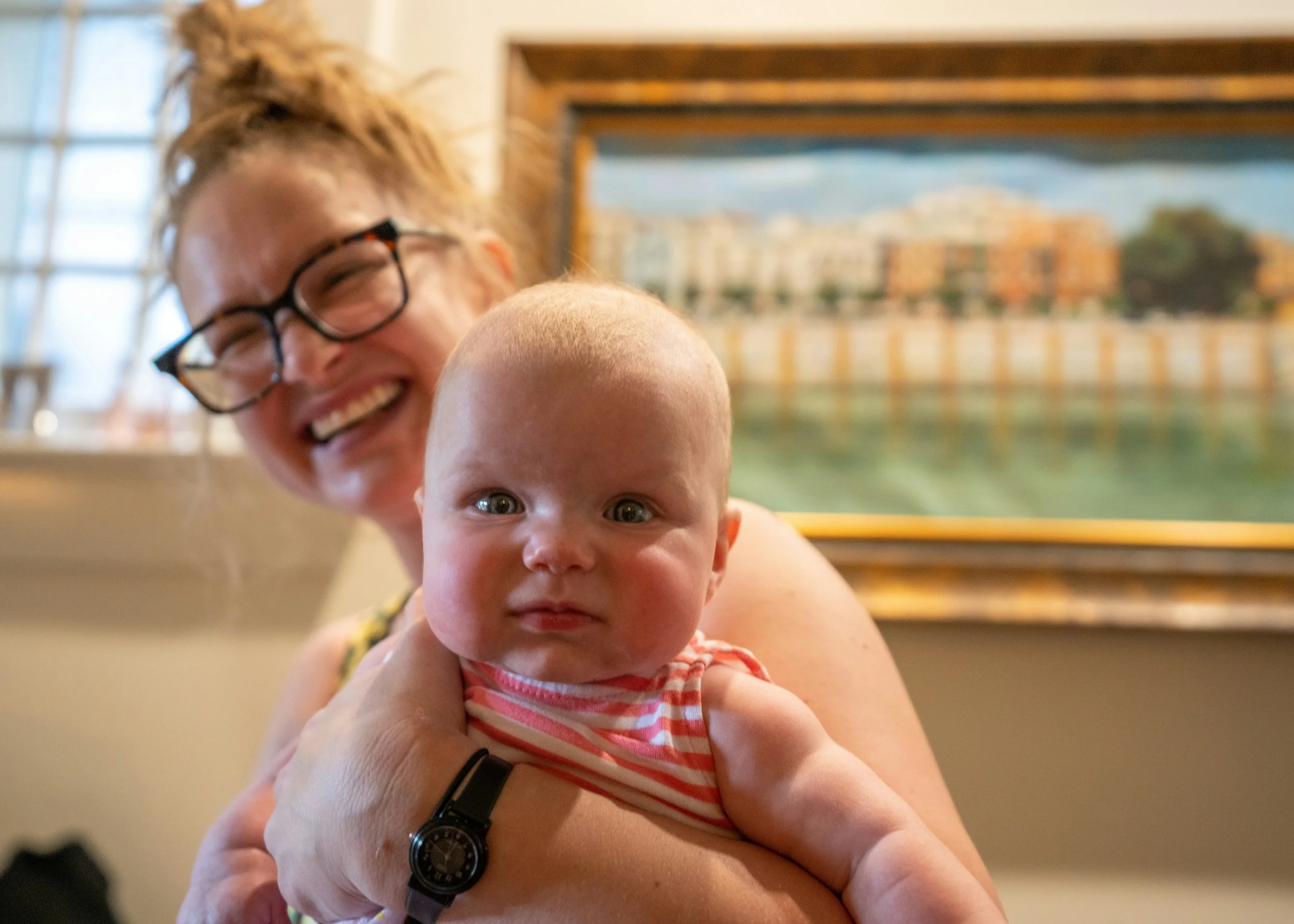 A young woman with glasses and curly hair smiling while holding a baby with big eyes, both indoors near a swimming pool window background.