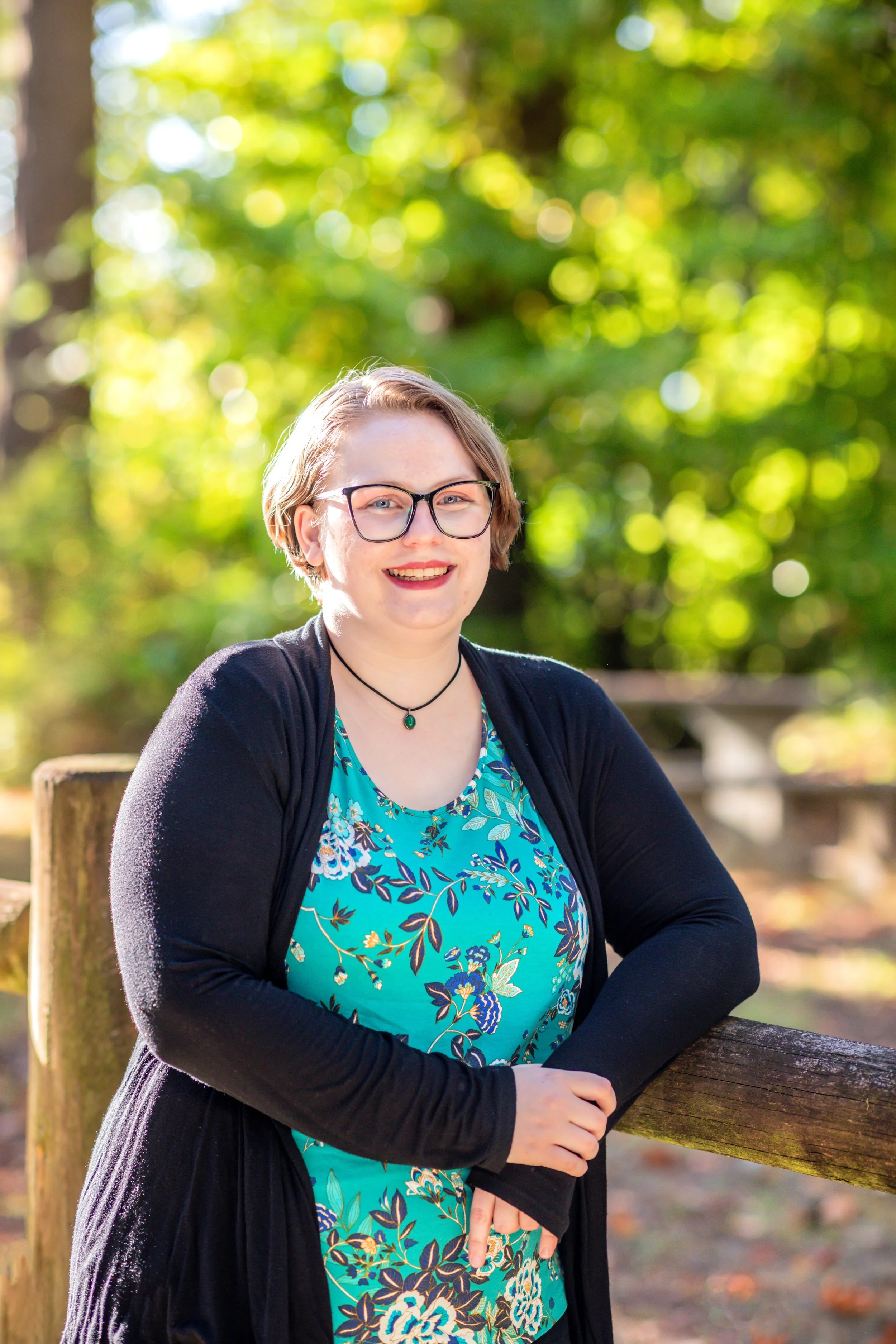 A young woman with short blonde hair, glasses, and a black necklace, wearing a turquoise floral dress and black cardigan, smiling outdoors in a park or wooded area.