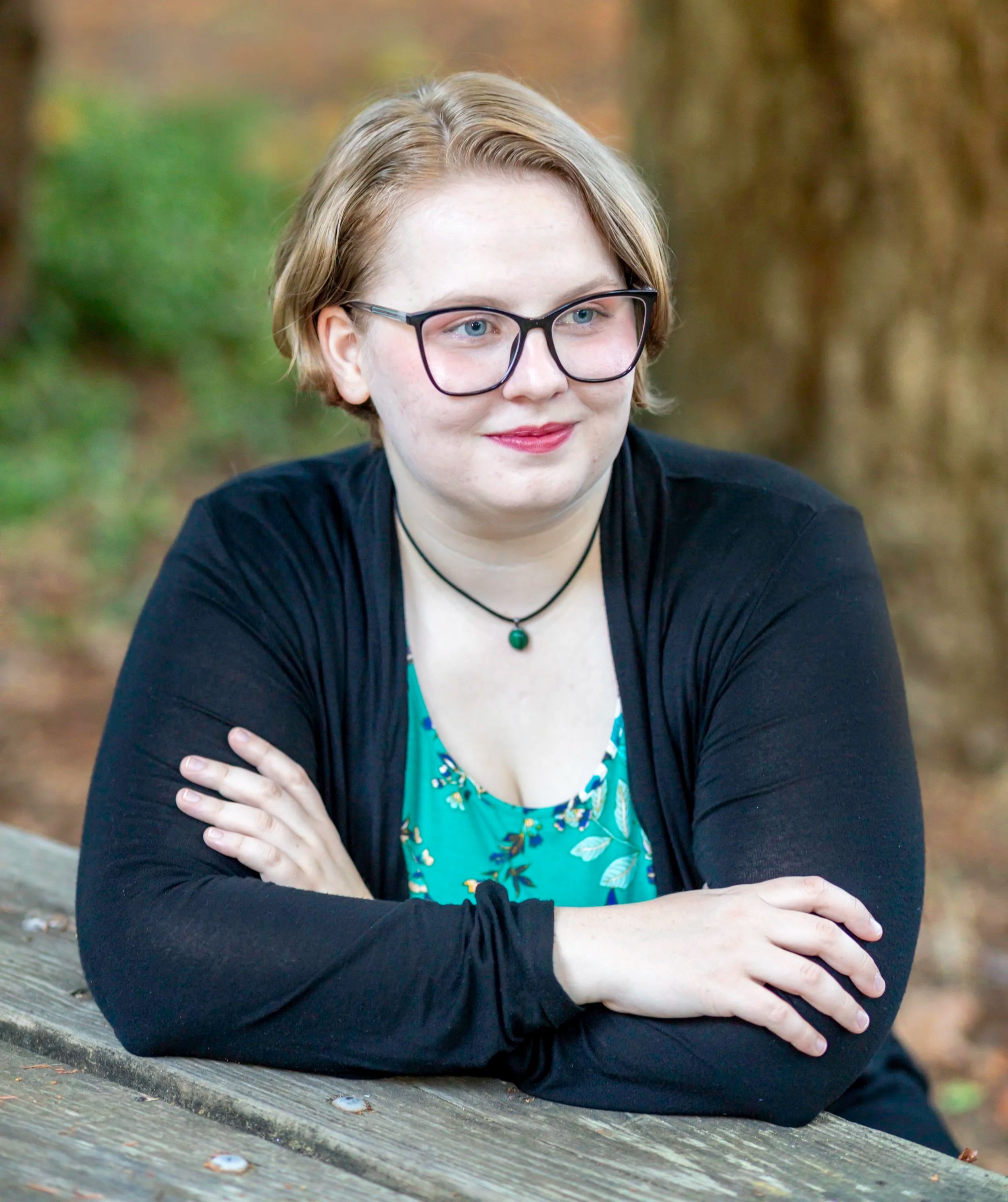A woman with short blonde hair, glasses, and a black shirt sitting outdoors on a wooden bench with her arms crossed and smiling slightly. The background is blurred with autumn foliage.