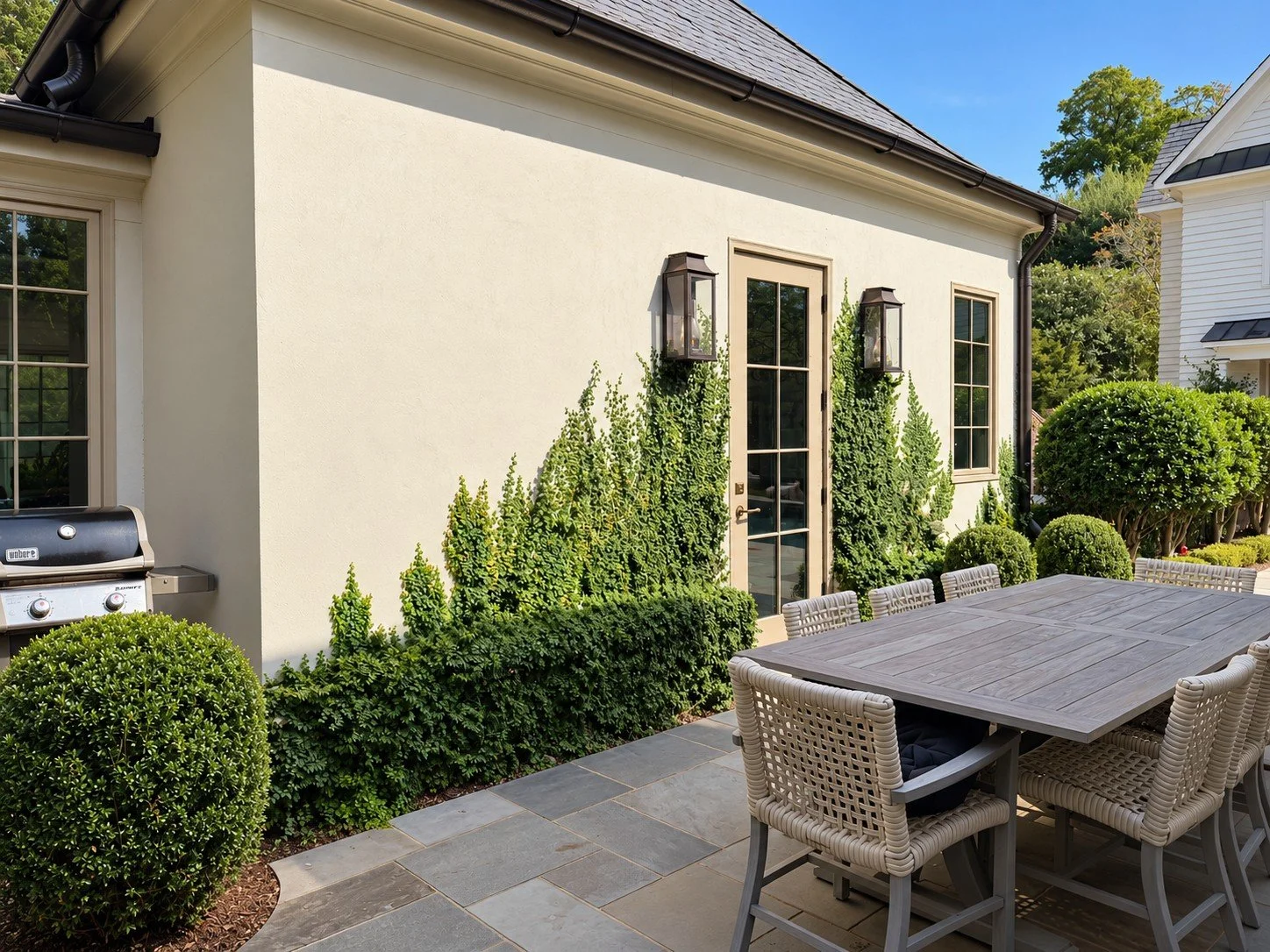 Outdoor dining patio with a wooden table, woven chairs and bluestone paving, surrounded by manicured evergreen hedges and climbing greenery that partially conceal a stainless steel grill beside the house.