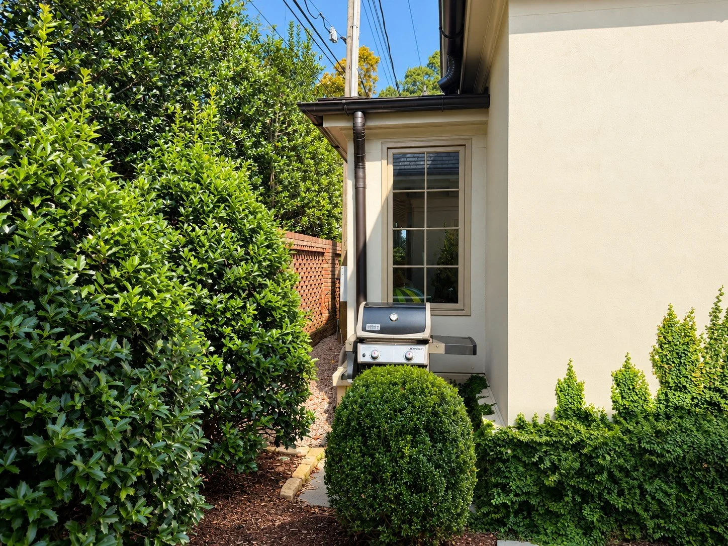 Stainless steel outdoor grill tucked into a discreet space beside the house, screened from the dining patio by tall evergreen hedges and rounded boxwoods while remaining easily accessible.