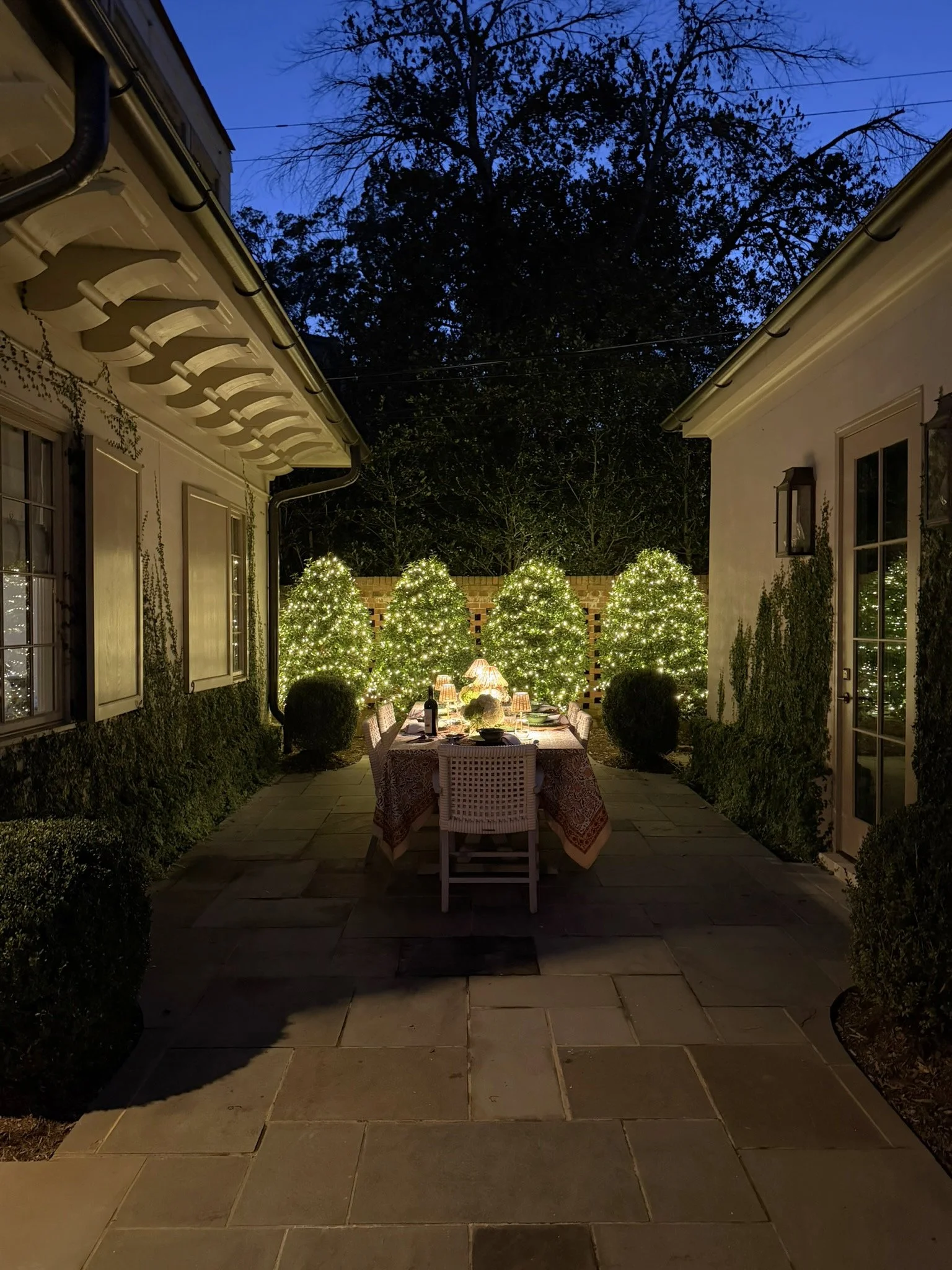 Intimate outdoor dining courtyard at night, framed by two buildings with climbing greenery, softly illuminated evergreen trees, a beautifully set dining table and warm lamps creating a welcoming garden retreat.