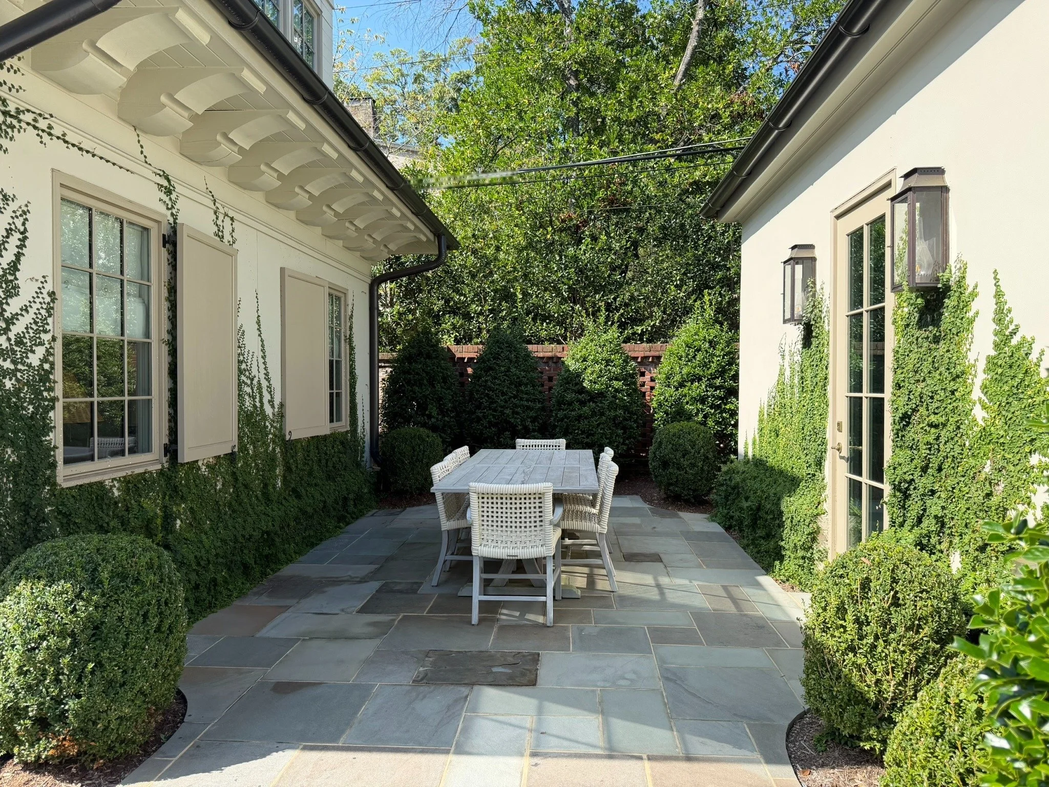 Secluded outdoor dining patio nestled between two buildings, with a wooden table, woven chairs, bluestone paving, creeping fig vines, evergreen hedges and rounded boxwoods creating an intimate garden retreat.
