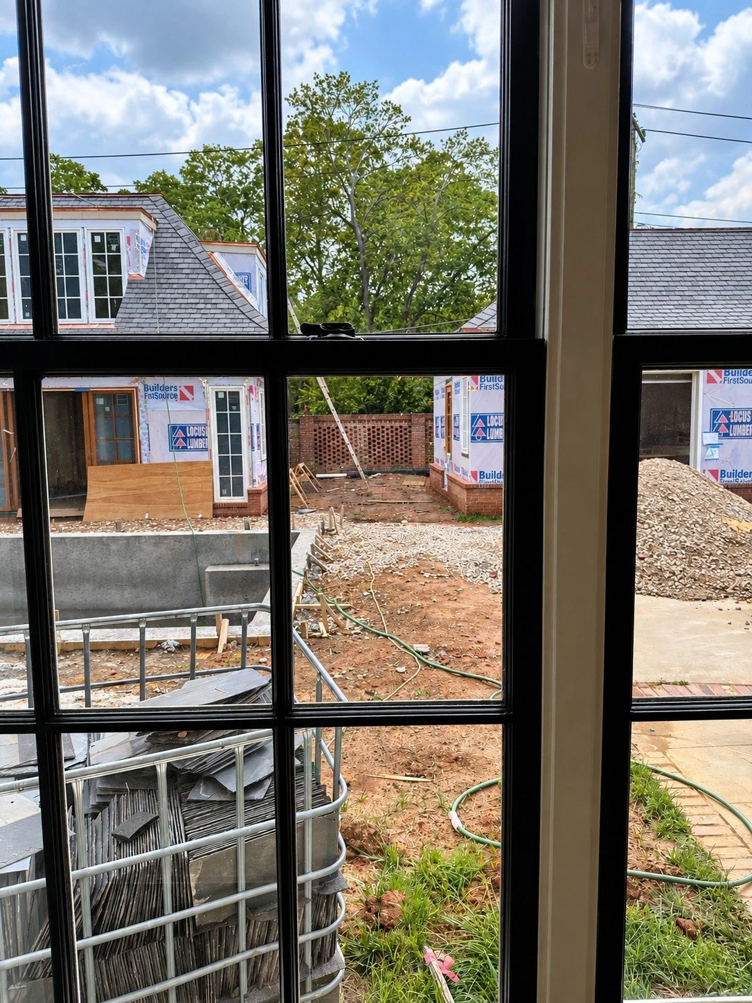 View through a black-framed window during backyard construction, showing unfinished pool and garden areas, exposed soil, building materials and two partially completed structures.