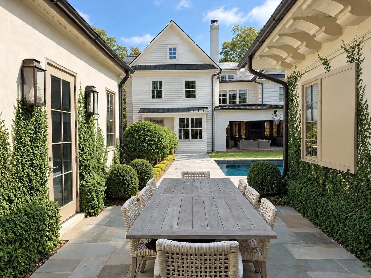 View from a secluded outdoor dining patio toward the main house and swimming pool, framed by two buildings, layered evergreen plantings, climbing vines and a long wooden dining table.