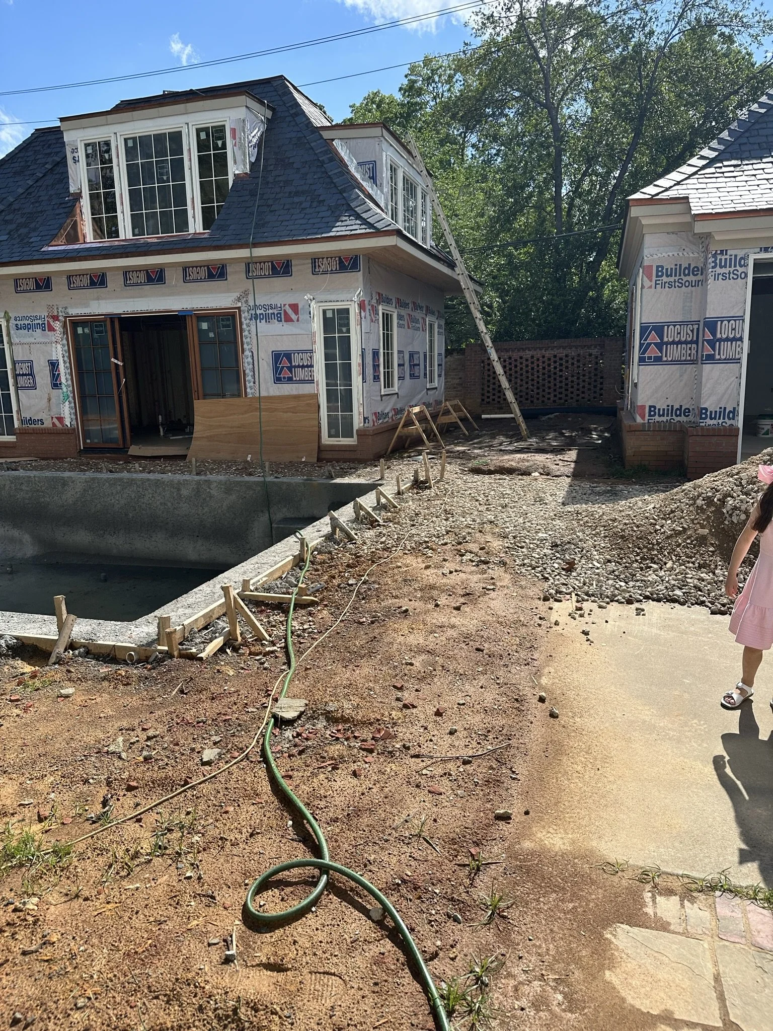 Outdoor space between two buildings during construction, showing unfinished ground, exposed building materials, a brick garden wall and the beginnings of a swimming pool.
