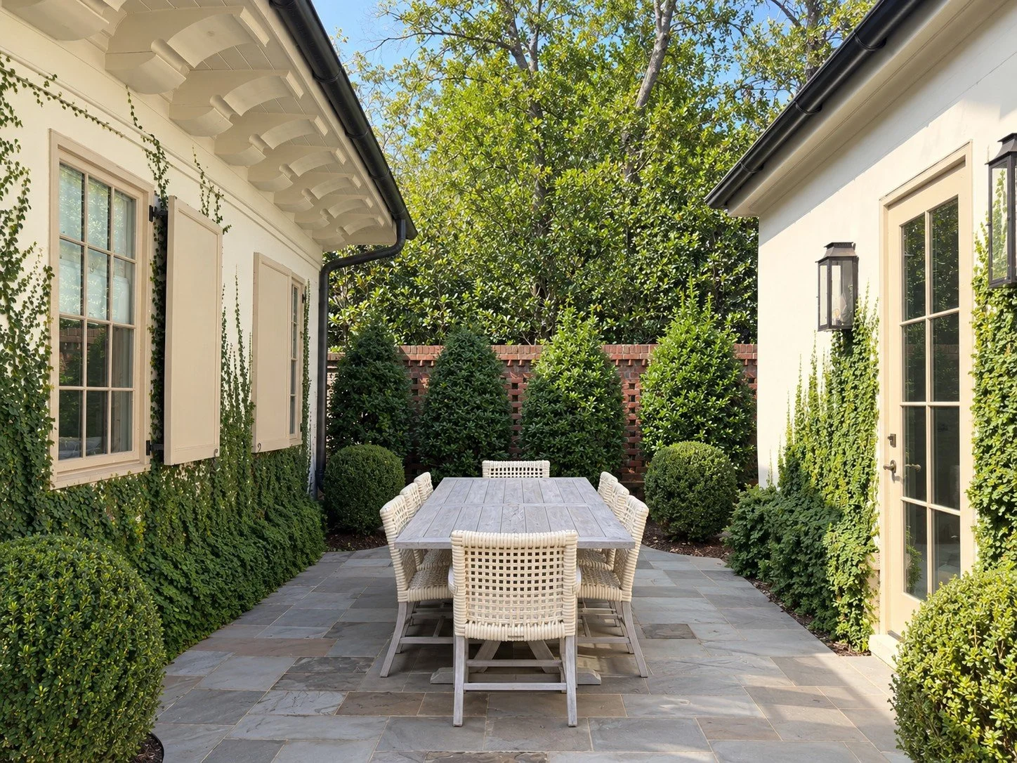 Intimate outdoor dining patio framed by two buildings, with a long wooden table, woven chairs, bluestone paving, layered evergreen hedges and climbing greenery creating a sheltered garden room.
