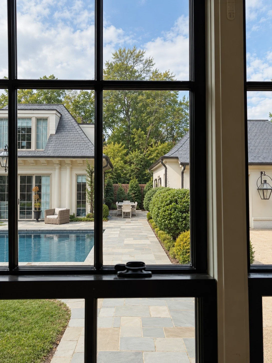 View through a black-framed interior window overlooking a landscaped courtyard with a swimming pool, bluestone walkway, white garden buildings, layered evergreen hedges and a secluded outdoor dining area.