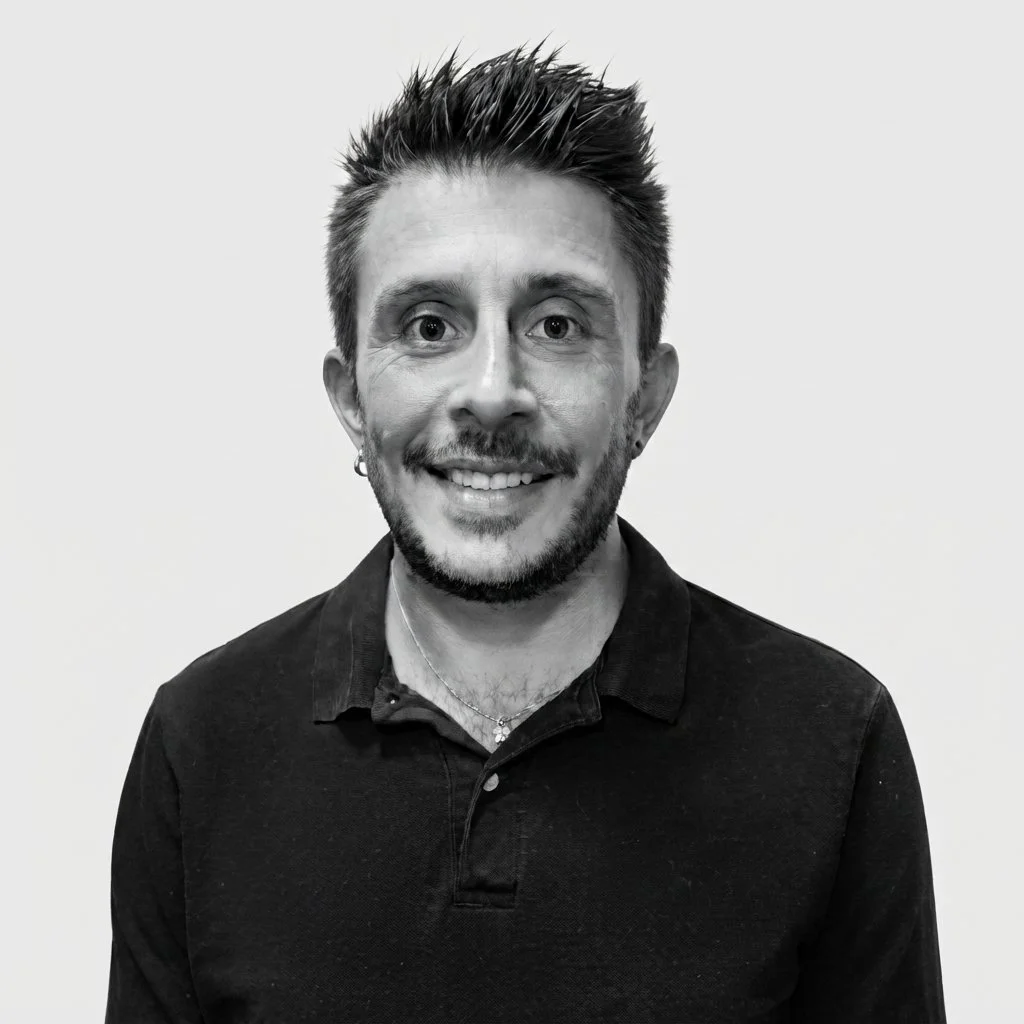Portrait d'un homme souriant, portant un polo noir, avec une barbe, des cheveux en pique, portant une boucle d'oreille et une chaîne