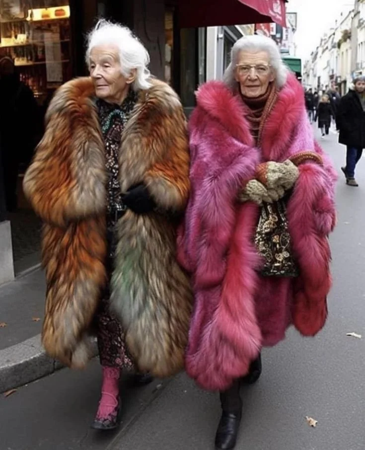 Two elderly women walking on a city sidewalk, wearing colorful fur coats—one in orange and brown, the other in pink.