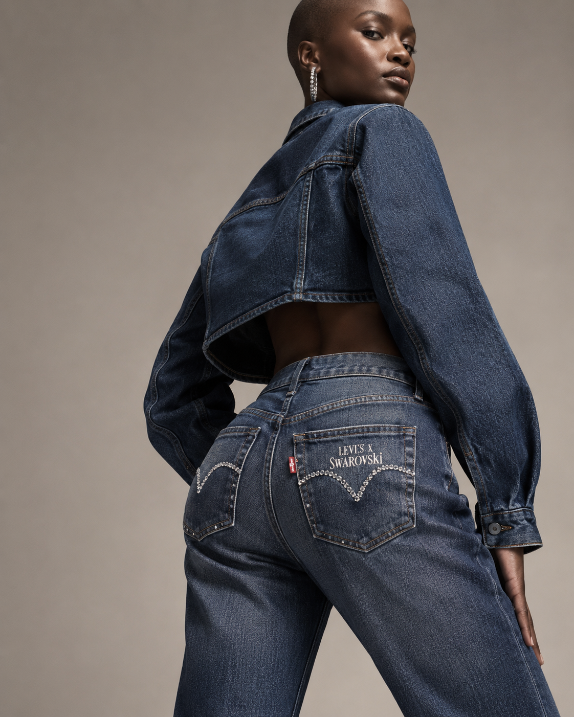 A woman with dark skin and a shaved head wearing a cropped denim jacket, high-waisted jeans with Swarovski rhinestone accents, and earrings, posing against a plain background.