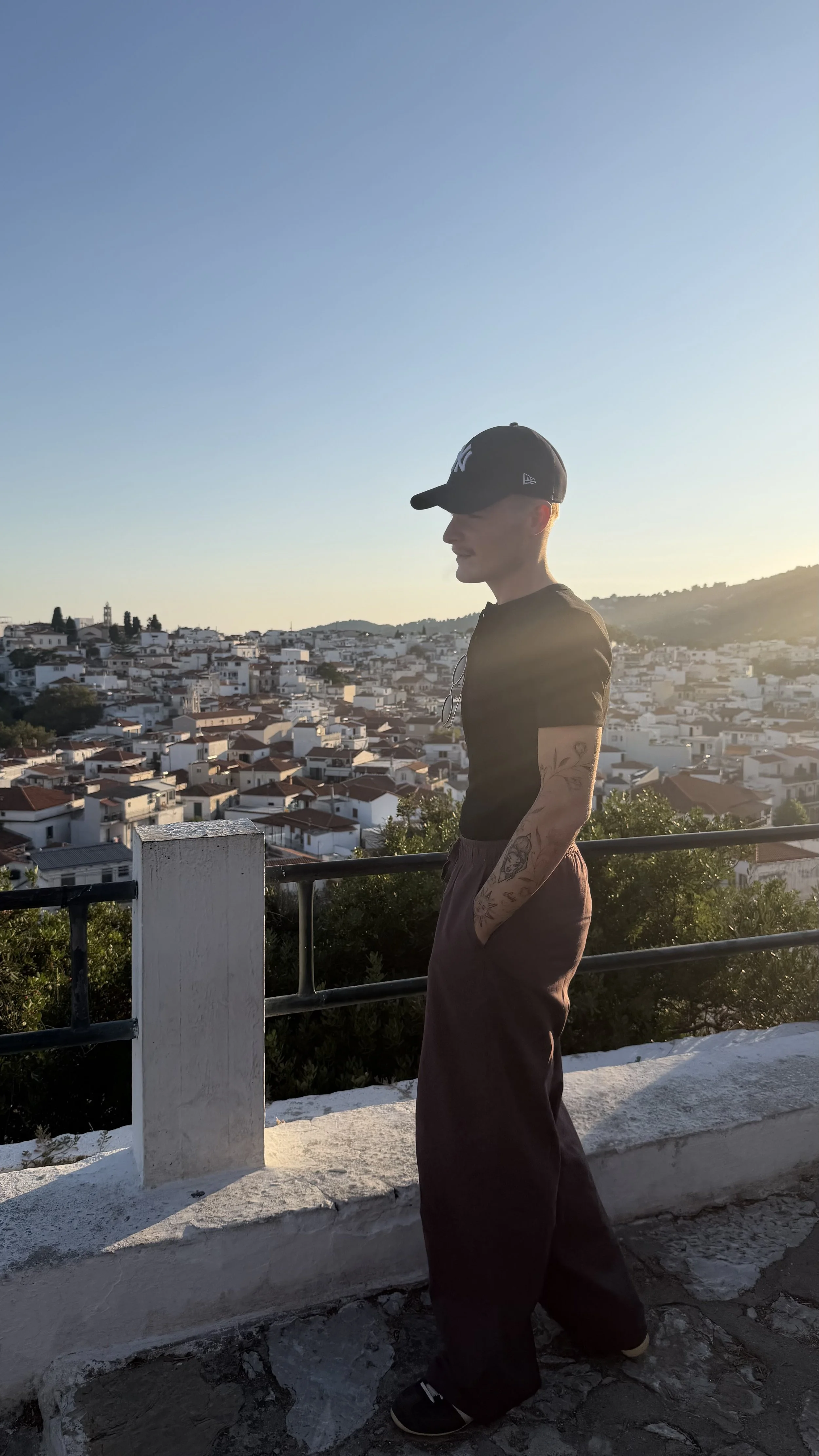 A young man wearing a black New York Yankees baseball cap, black t-shirt, and dark pants stands with hands in pockets on a balcony overlooking a town with white houses during late afternoon or early evening sunset.