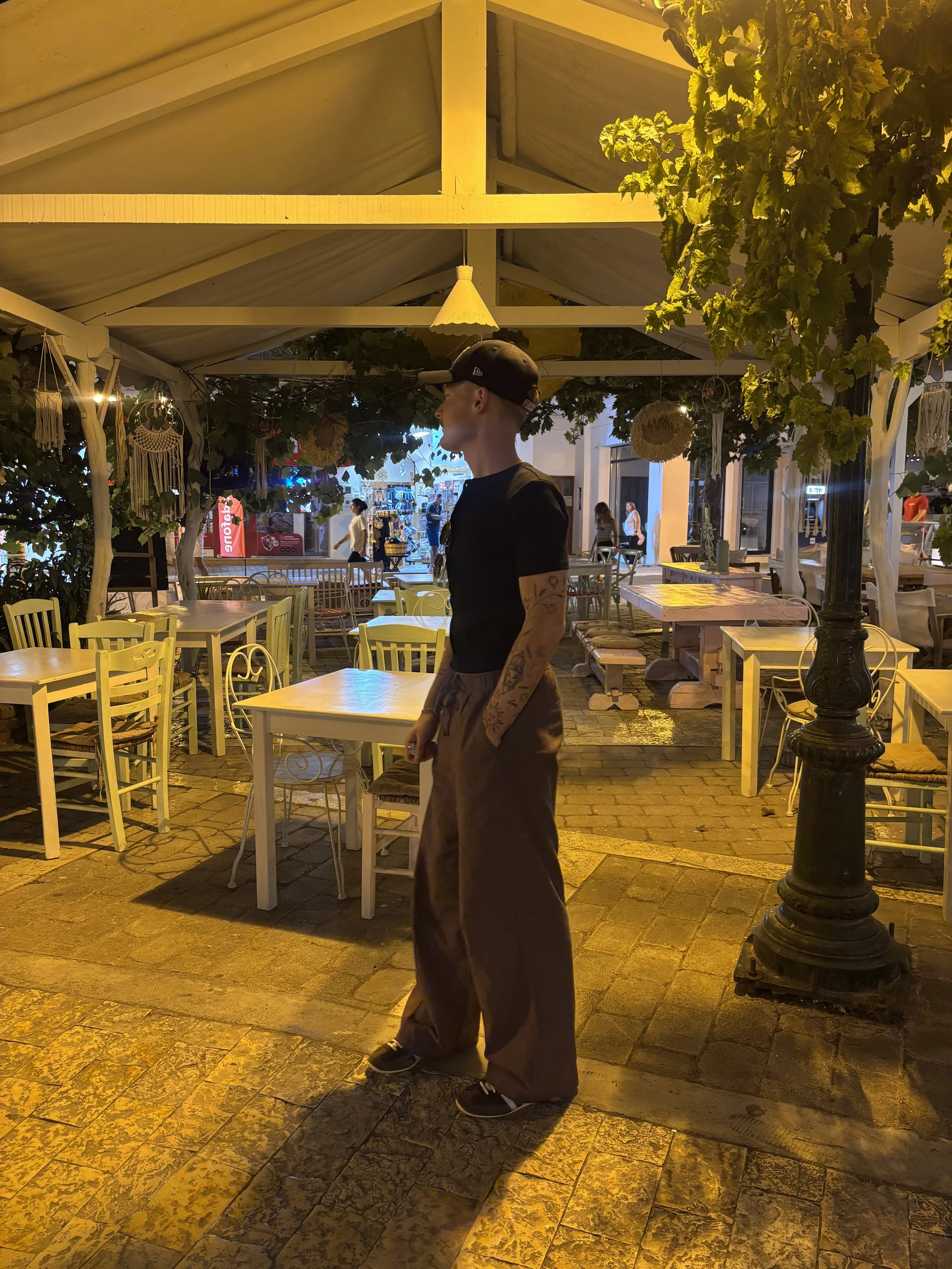 A young man wearing a black baseball cap, black t-shirt, brown baggy pants, and sneakers standing in an outdoor seating area of a restaurant or cafe at night. The area is decorated with hanging dreamcatchers and plants, with yellow lighting, and there are people walking and shopping outside in the background.
