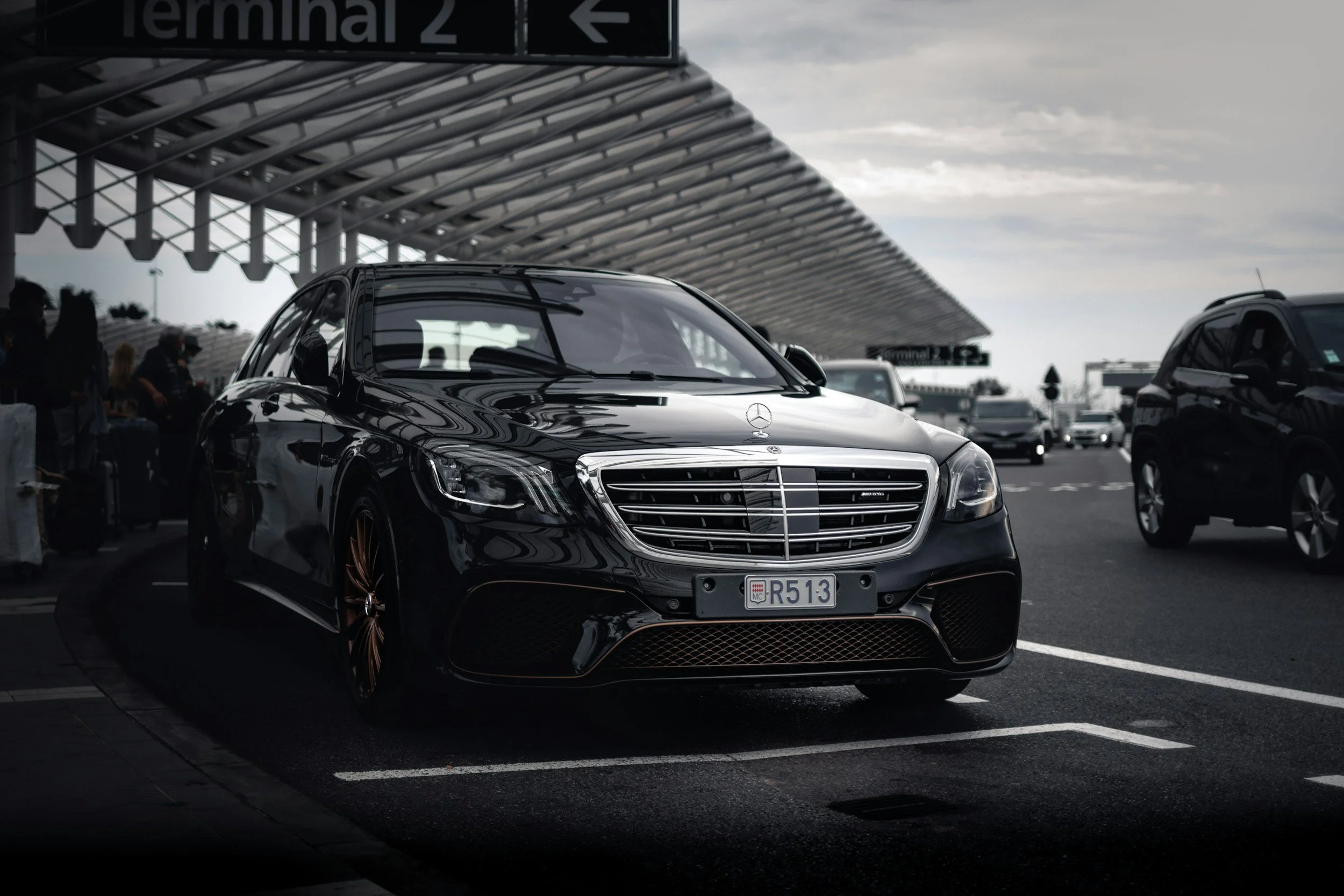 A black luxury Mercedes-Benz car parked at an airport terminal, with terminal 2 signs overhead and other vehicles in the background.