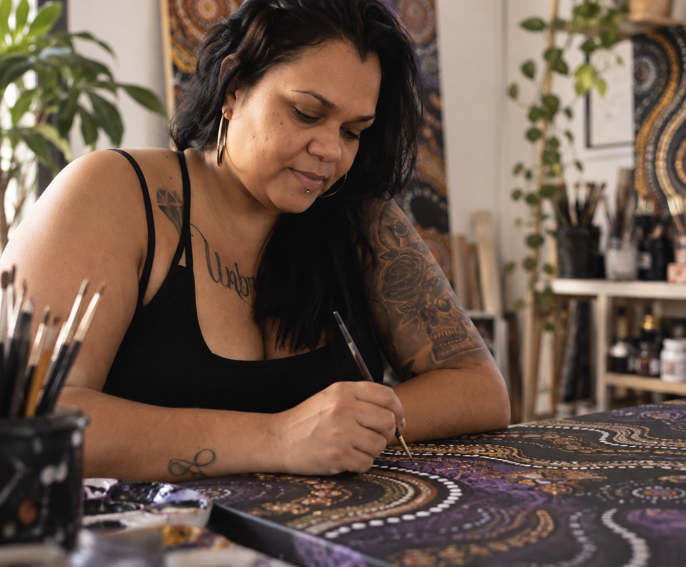 A woman with black hair and tattoos painting a detailed, colorful Aboriginal Australian design on a canvas.