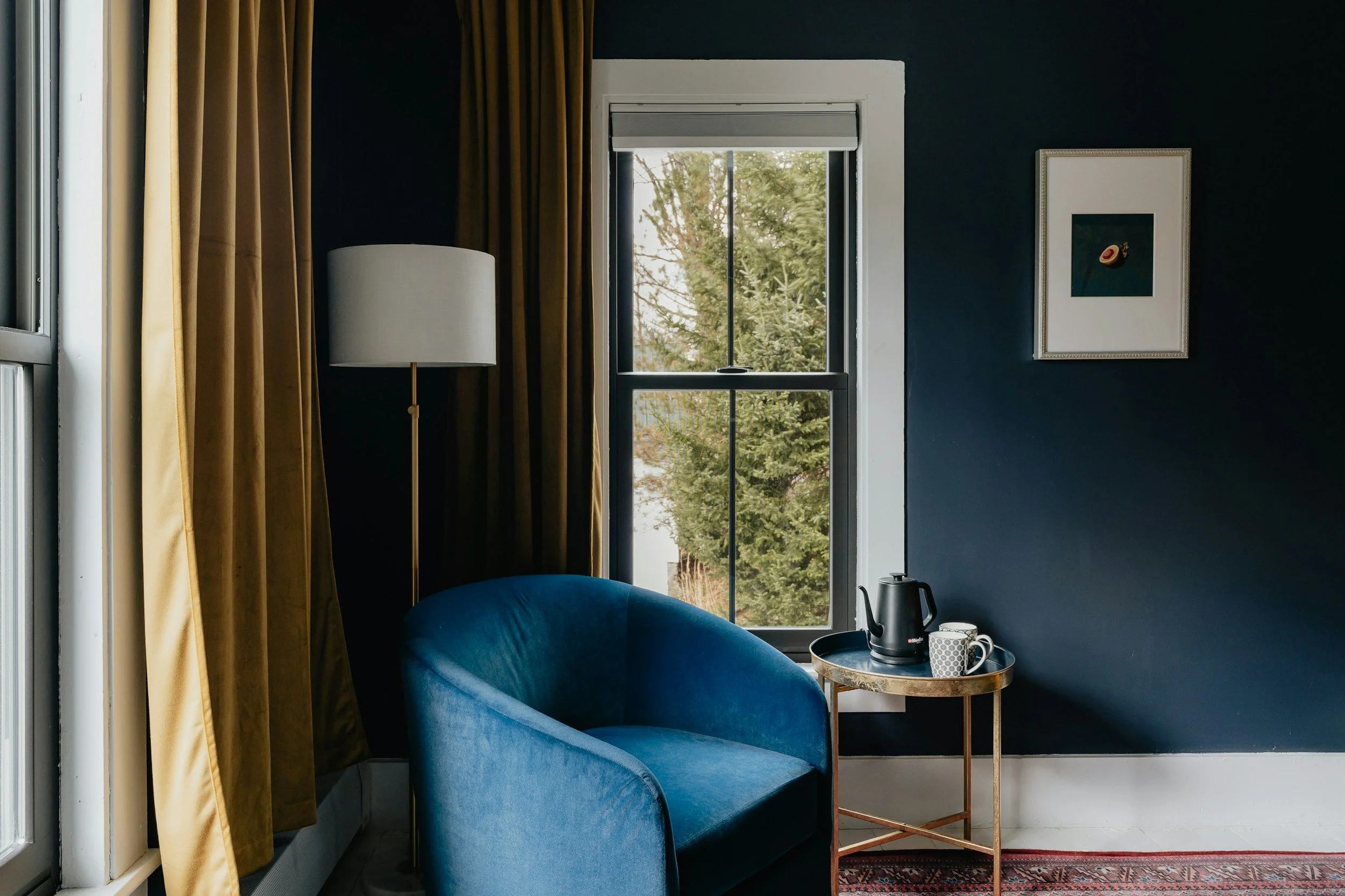 A cozy corner of a room with a blue armchair, a wooden side table with a black kettle, a white mug, and a small decorated bowl. There is a window with white trim and dark curtains, and a framed picture on the black wall. Outside the window, trees are visible.