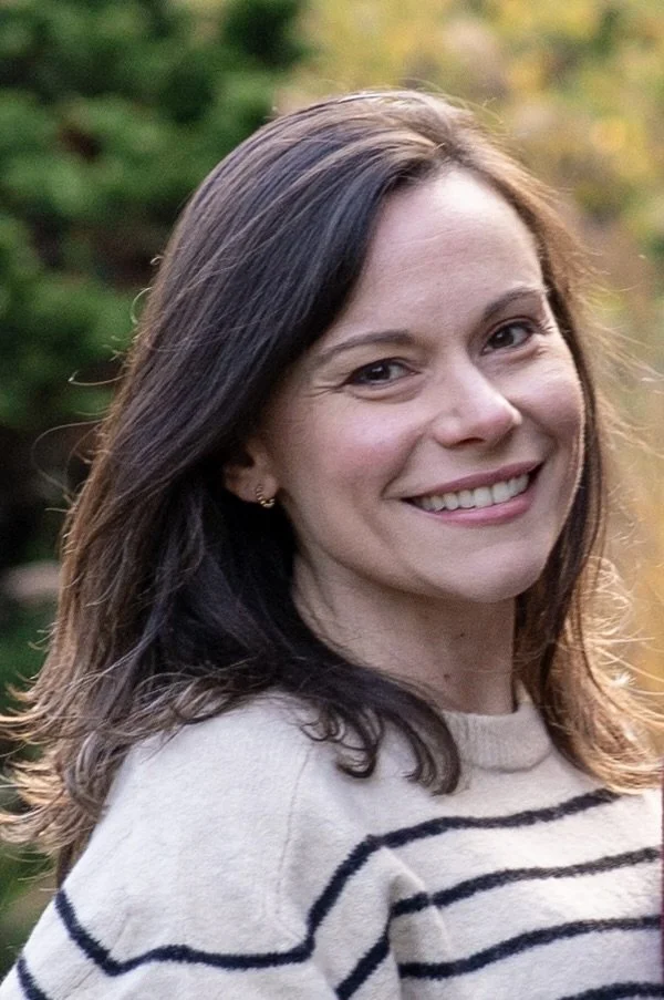 A woman with brown hair and a big smile, wearing a beige and black striped sweater, outdoors with blurred green foliage in the background.
