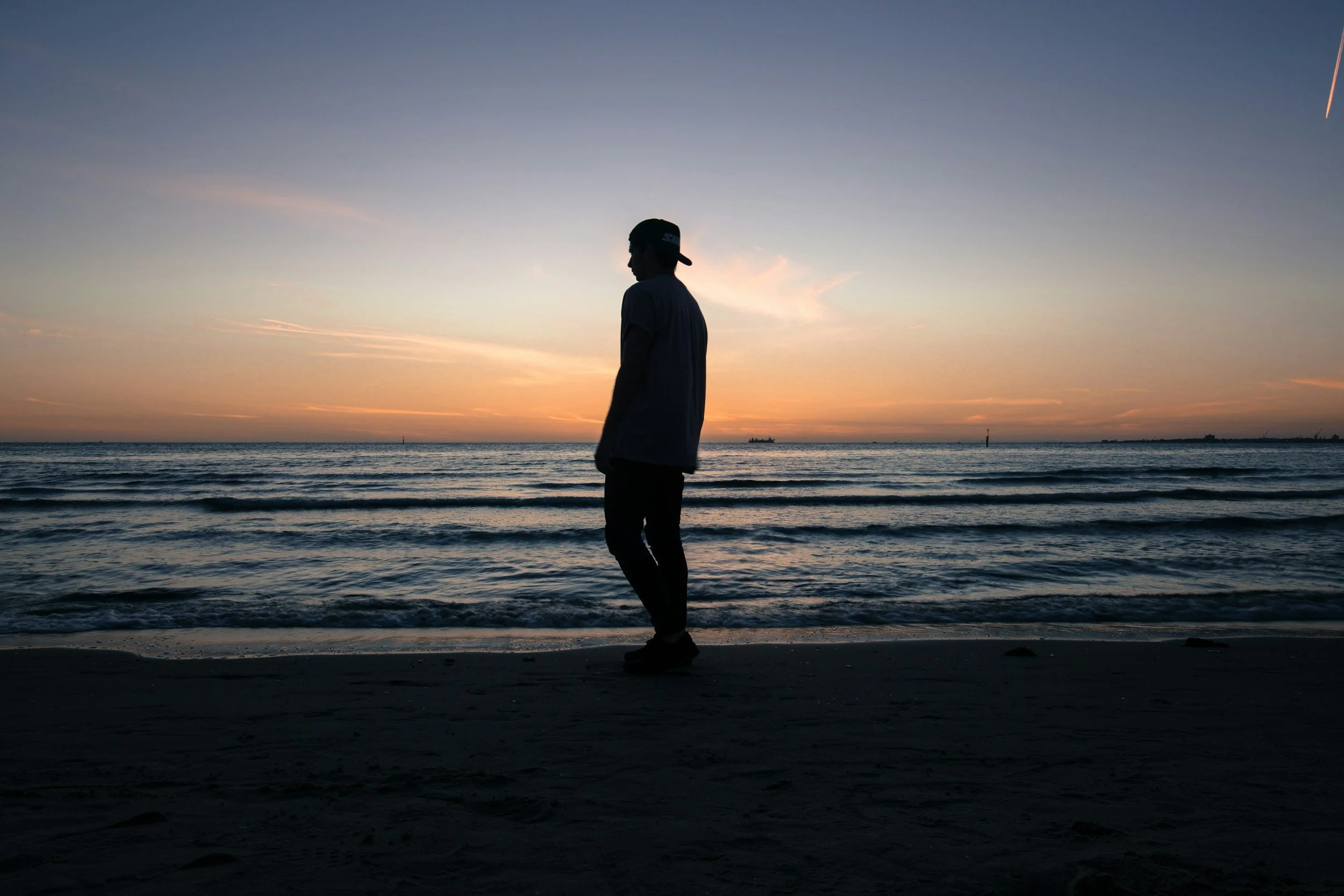 Silhouette of a person wearing a cap walking along the beach at sunset, with calm waves and an orange sky in the background.