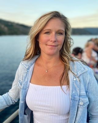 A woman with long blonde hair, wearing a light denim jacket over a white top, standing outdoors near a body of water with hills in the background.