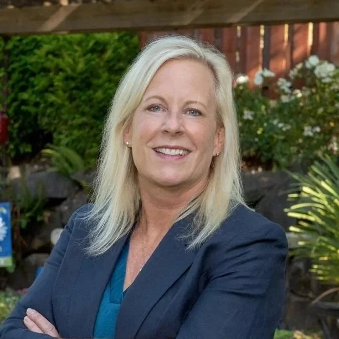 A smiling woman with shoulder-length blonde hair, wearing a navy blazer, standing outdoors in a garden with green plants and white flowers.