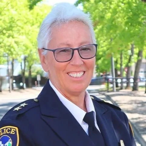 A smiling female police officer with short white hair, glasses, and a dark blue uniform, standing outdoors with green trees in the background.