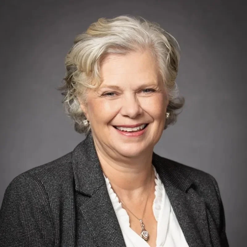 A smiling older woman with short, curly gray hair, wearing a dark blazer, white blouse, and pearl earrings, against a plain gray background.