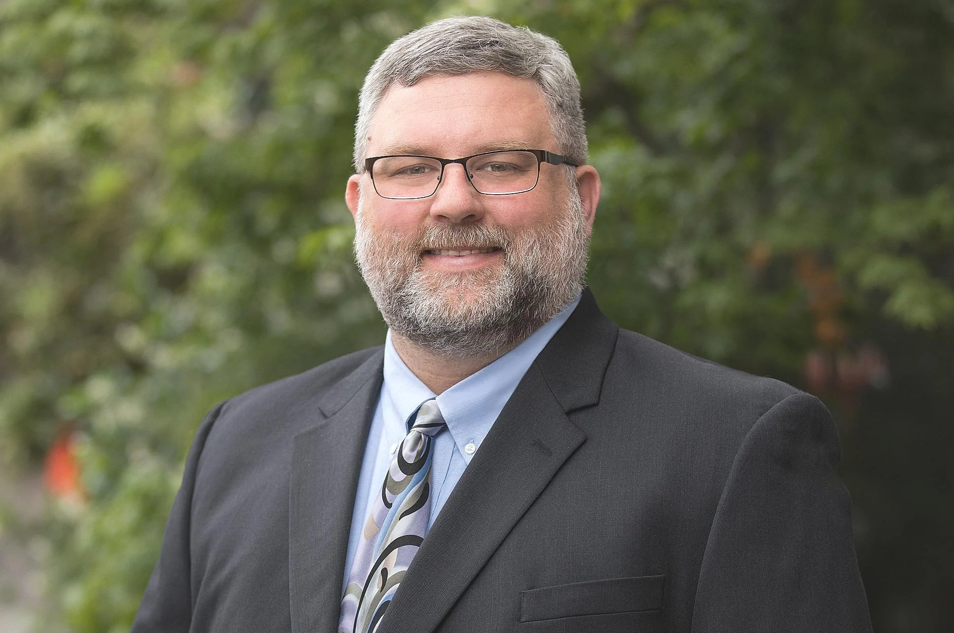 A middle-aged man with gray hair, glasses, and a beard, dressed in a black suit, light blue shirt, and patterned tie, standing outdoors with greenery in the background.