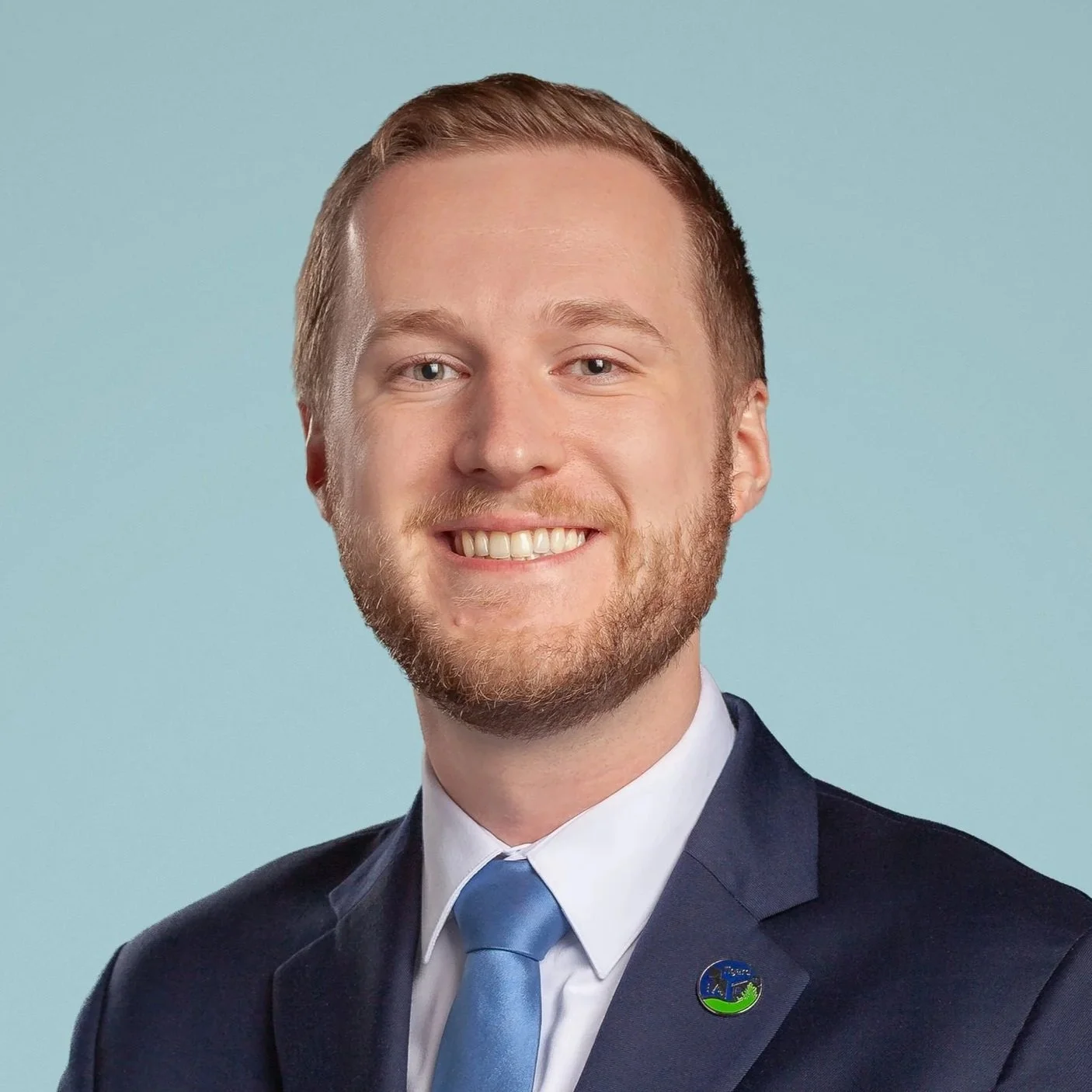 A young man with light skin and a beard wearing a dark suit, white shirt, and light blue tie, smiling in front of a light blue background.
