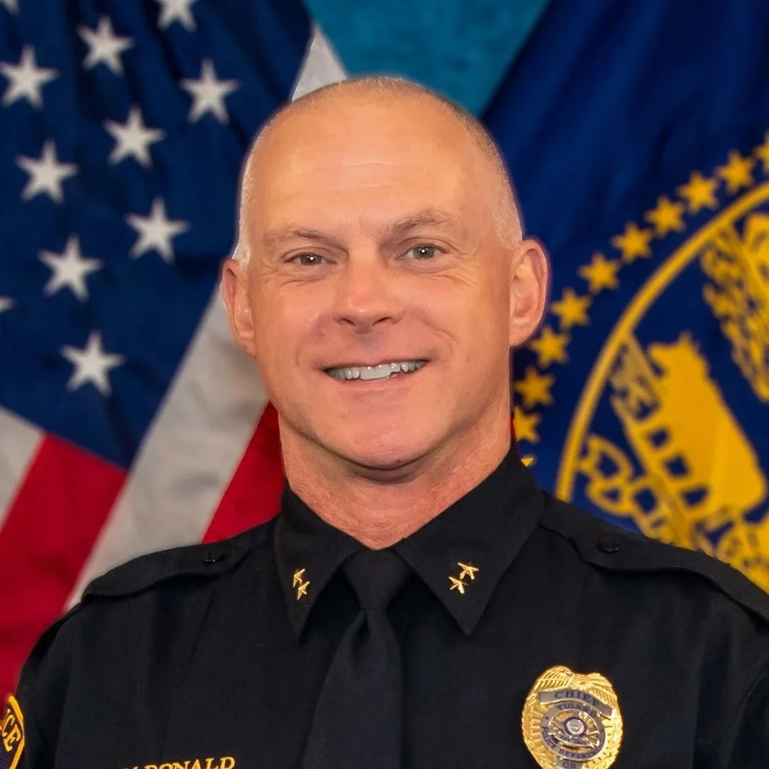 A smiling police officer in uniform with eagle insignia, standing in front of American and police flags.
