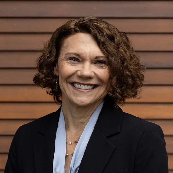 A smiling woman with curly brown hair wearing a black blazer and a light blue blouse, standing against a wooden slat background.