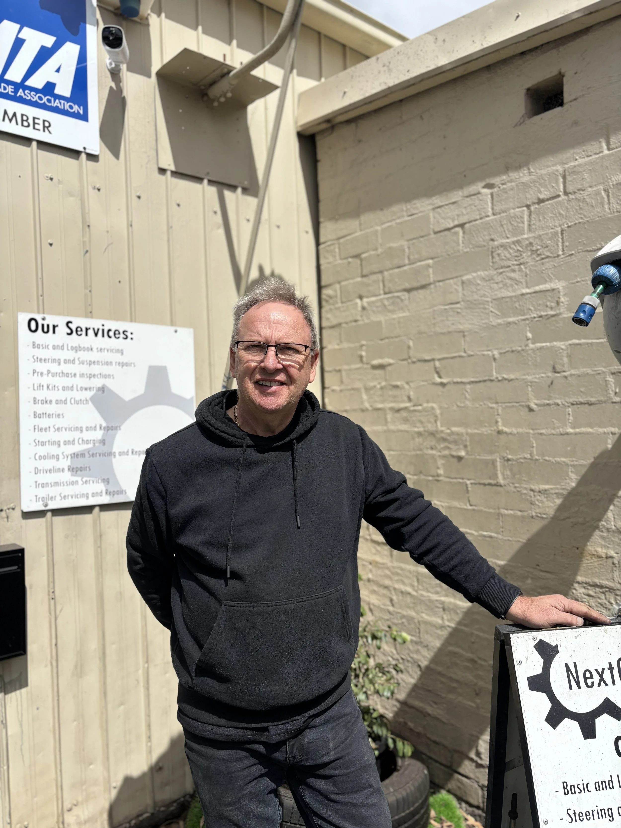 A man with glasses wearing a black hoodie standing outside near a beige brick wall and a wooden fence. He is smiling and has one hand resting on a black and white sign with words related to vehicle services. There is a security camera and an electrical box mounted on the wall, and part of a white sign with a list of services is visible in the background.