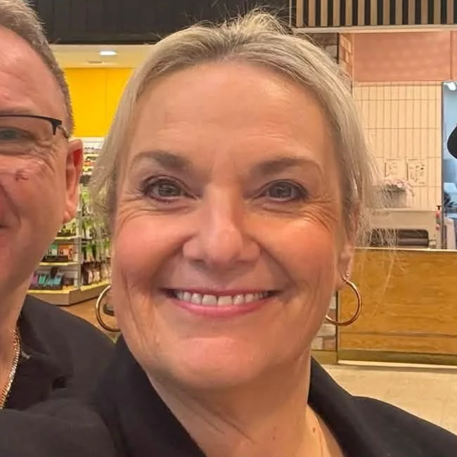 Close-up of a smiling woman with blonde hair, wearing hoop earrings, in a store or cafe environment.