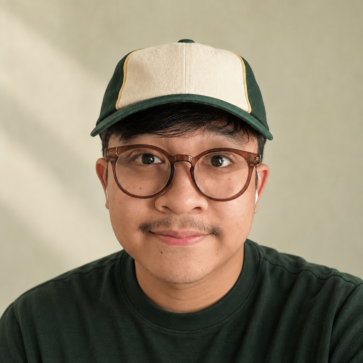 A young man with glasses and facial hair wearing a dark green shirt and a green and beige cap, smiling at the camera against a plain background.