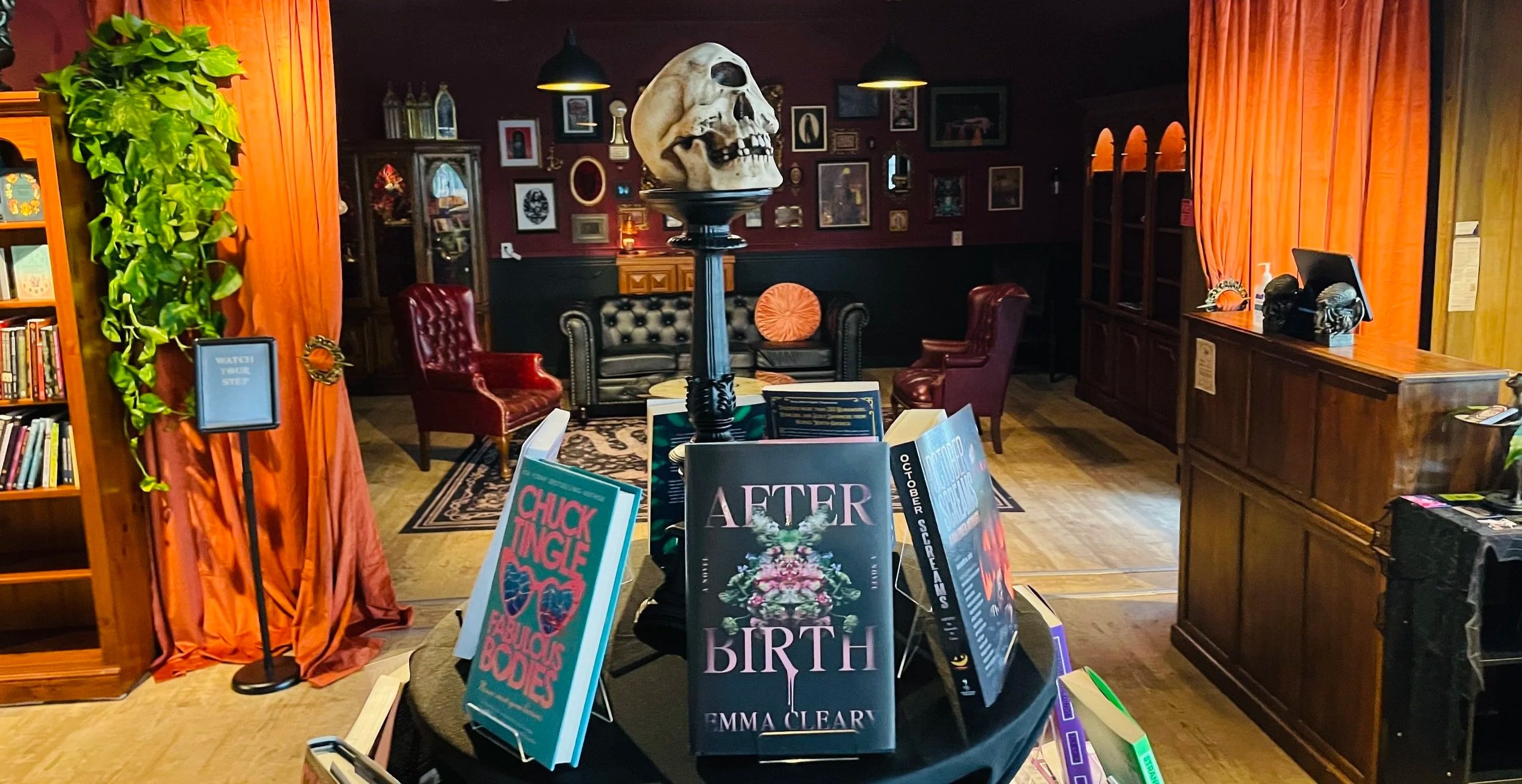Interior at Infernal Annex, in Olympia, Washington. Gothic and vintage decor throughout, including a large skull on a tall black pedestal in the center, framed pictures, bookshelves, and a table displaying books.