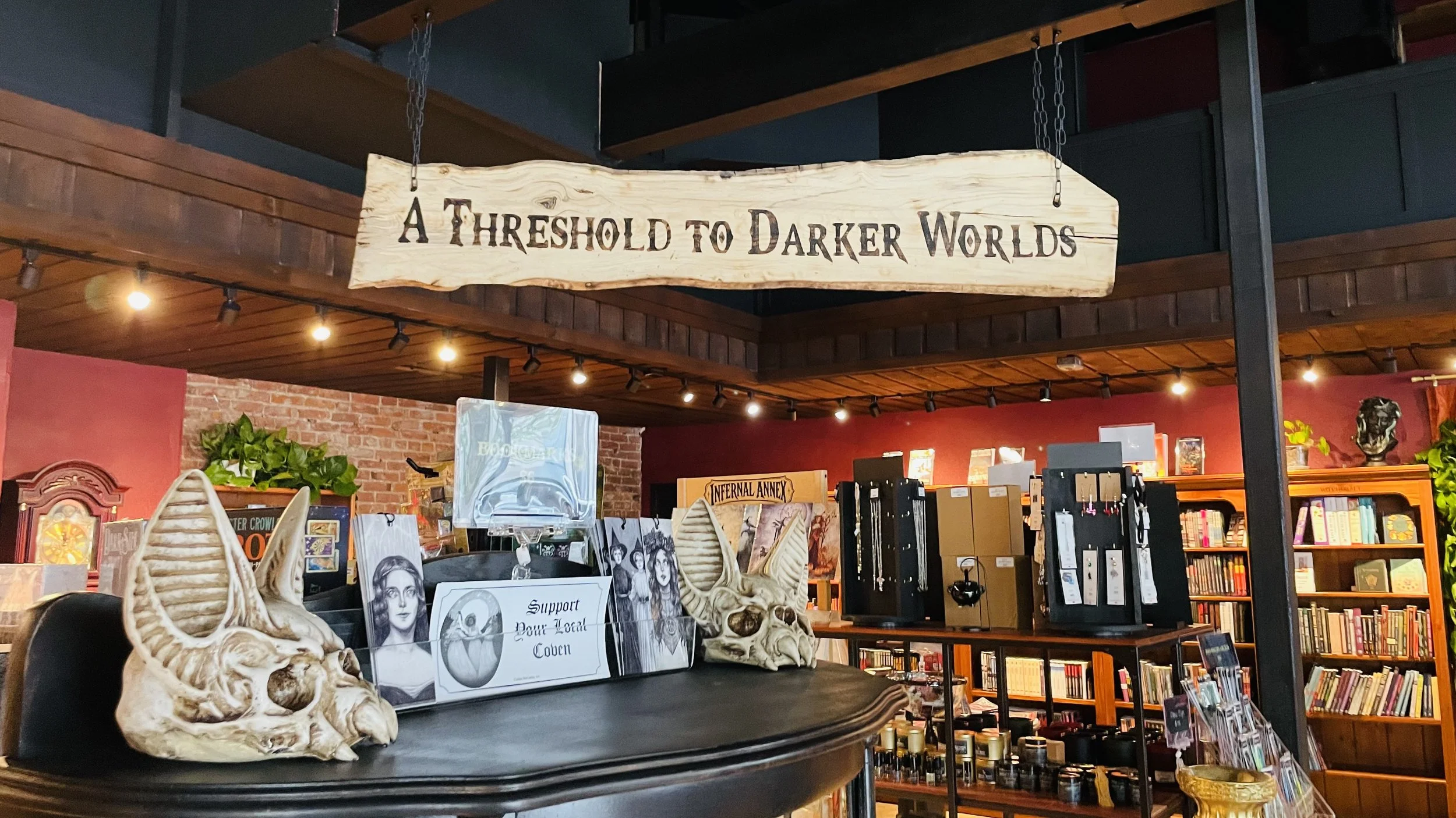 A wooden sign hangs from a chain that reads 'A Threshold to Darker Worlds' inside Olympia's latest horror book shop. Below the sign, there are skull sculptures and artwork, with shelves filled with books in the background.