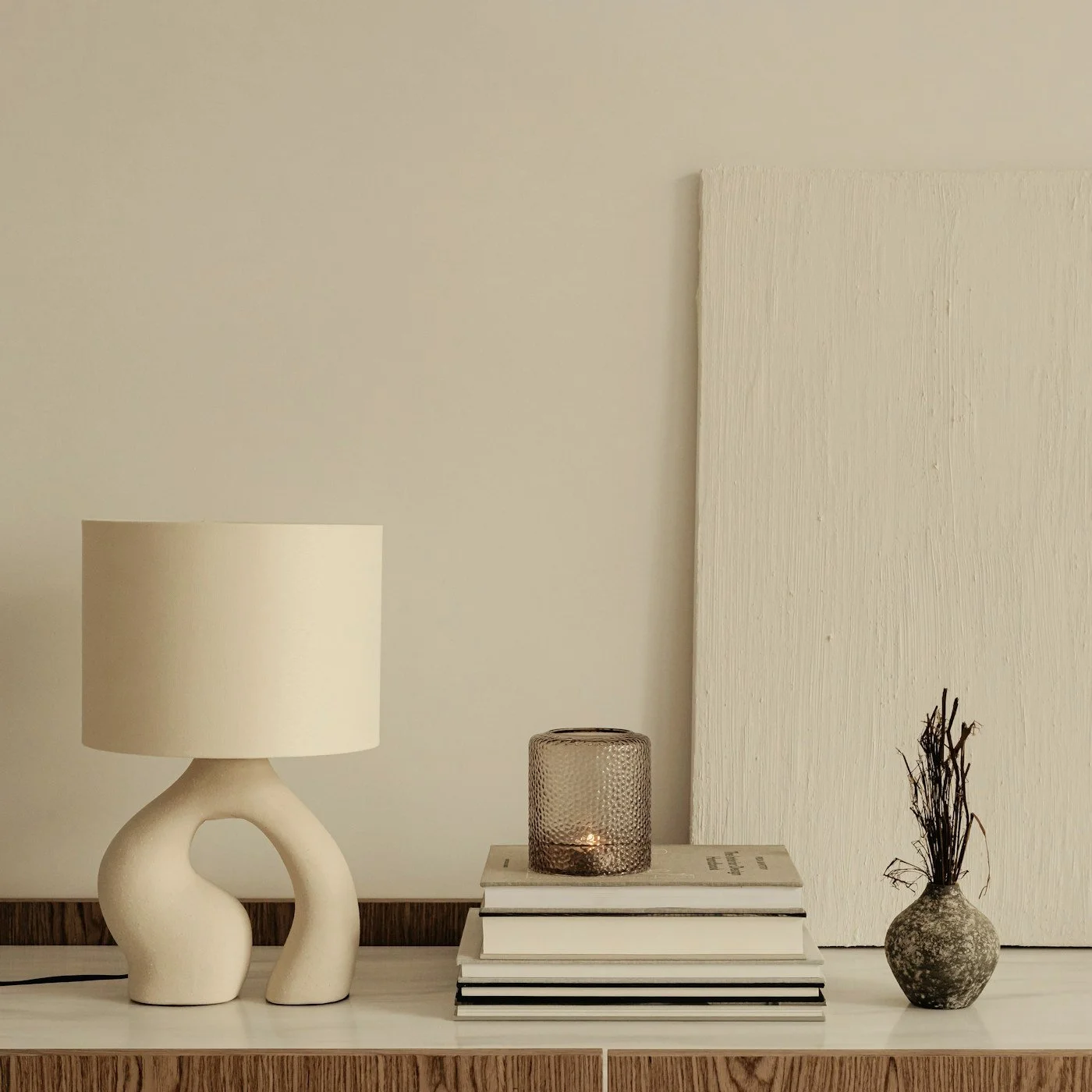 Neutral-toned minimalist console featuring an abstract ceramic table lamp, stacked books, textured candle holder, and dried branches in a vase.