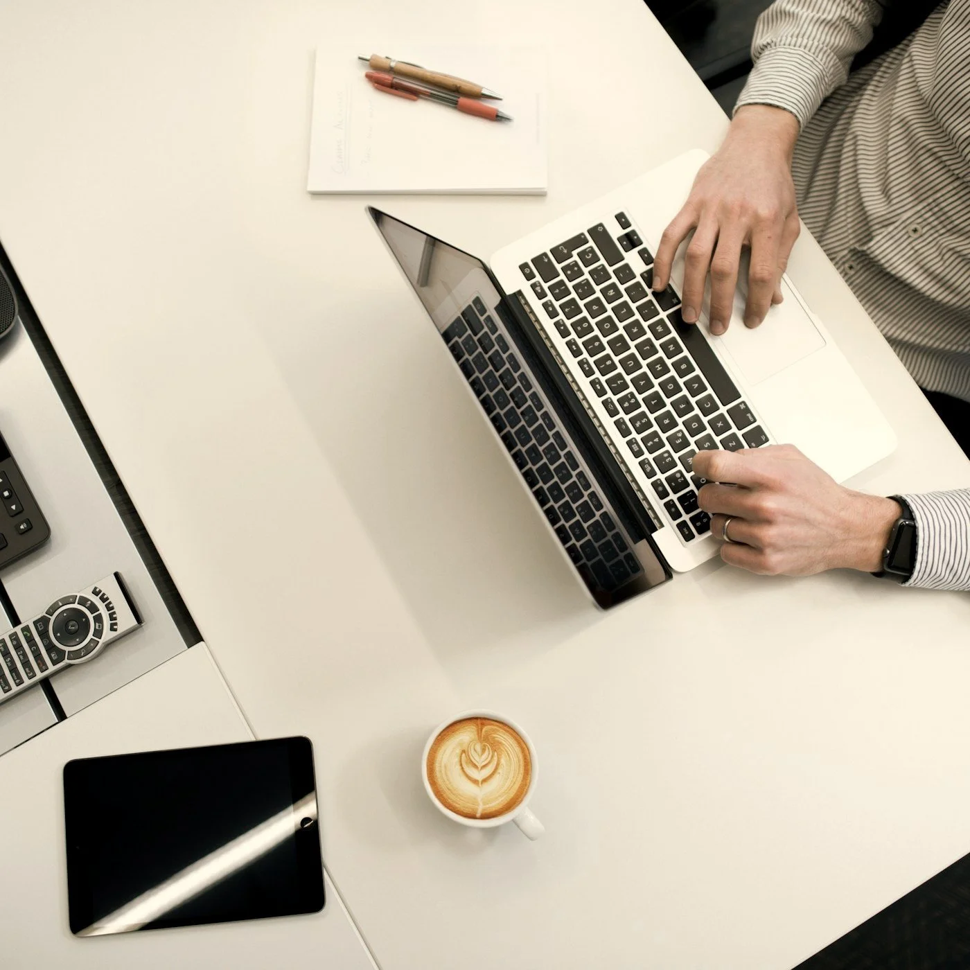Top-down view of a person working on a laptop at a desk with a notebook, pens, tablet, remote control, and latte.
