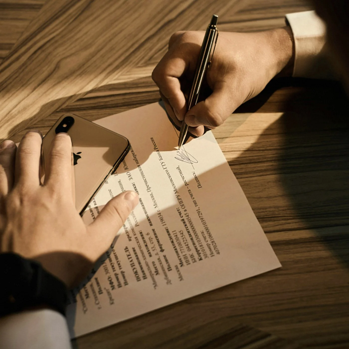 Person signing a document with a pen beside a smartphone on a wooden table