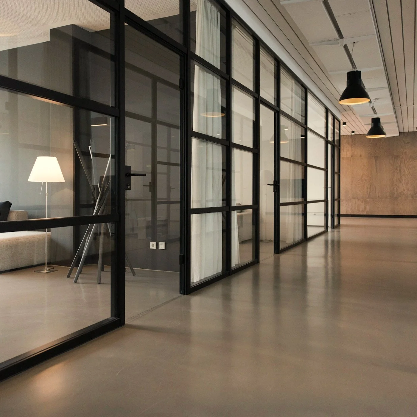 Modern office hallway with black-framed glass partitions, pendant lights, polished concrete floors, and a lounge area.