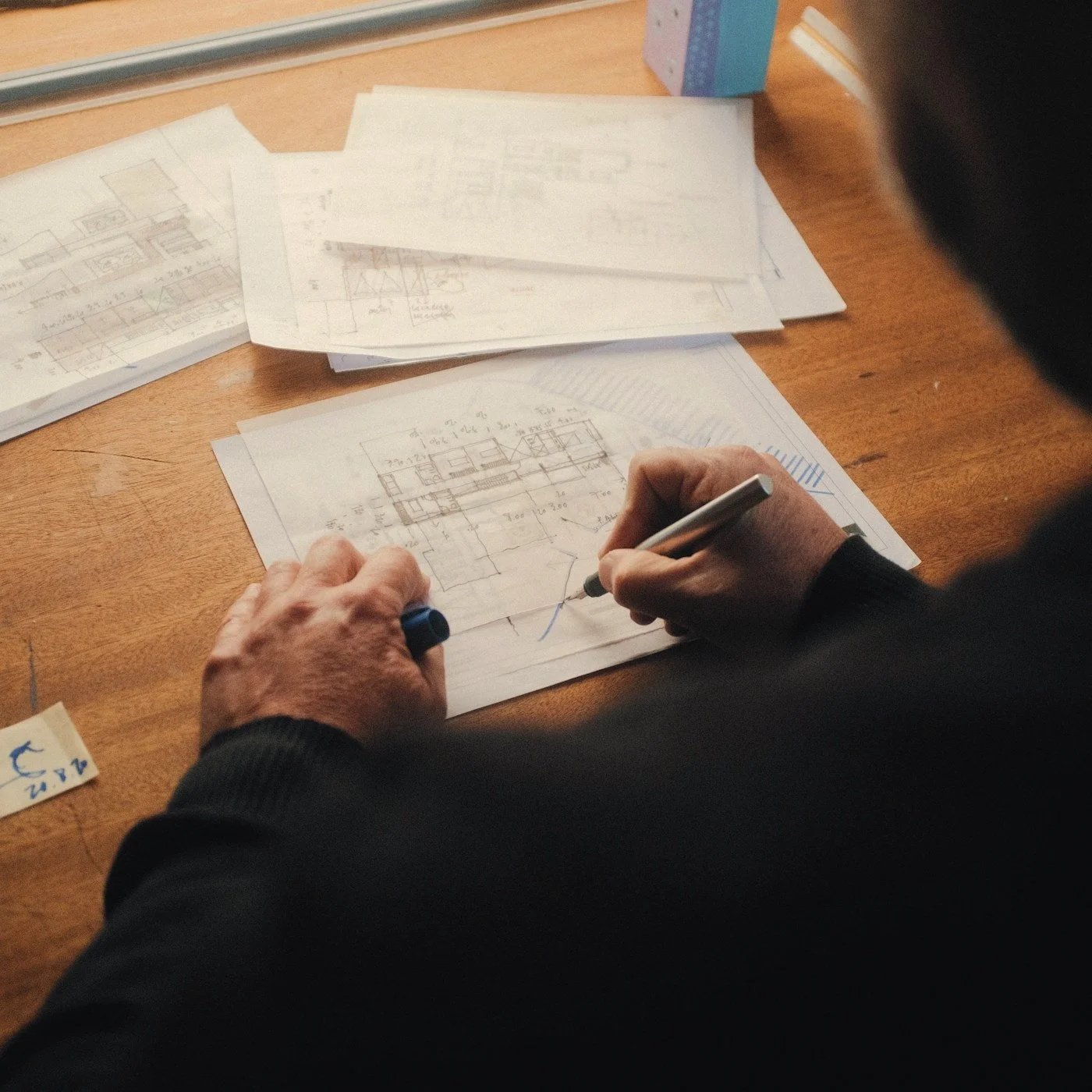 Person sketching architectural floor plans with a pen on paper at a wooden desk