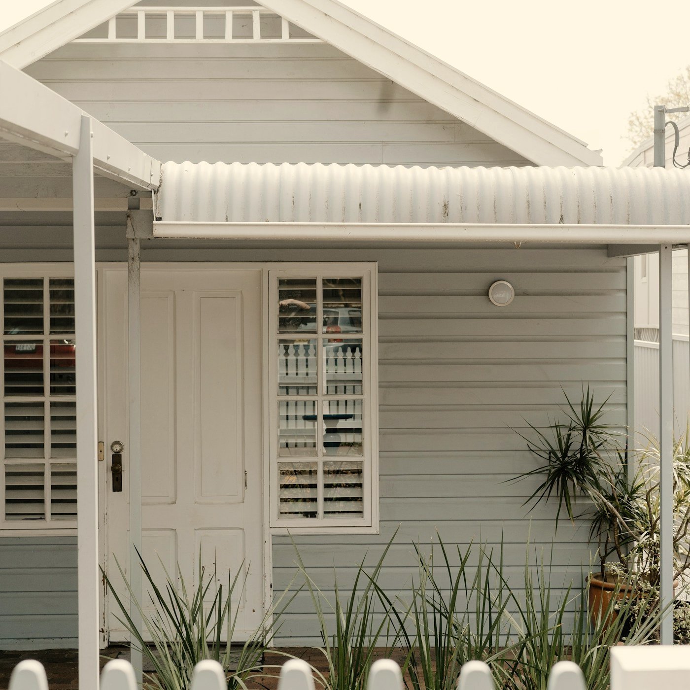 White cottage-style house entrance with a covered porch, paneled door, multi-pane windows, corrugated awning, and potted plants.
