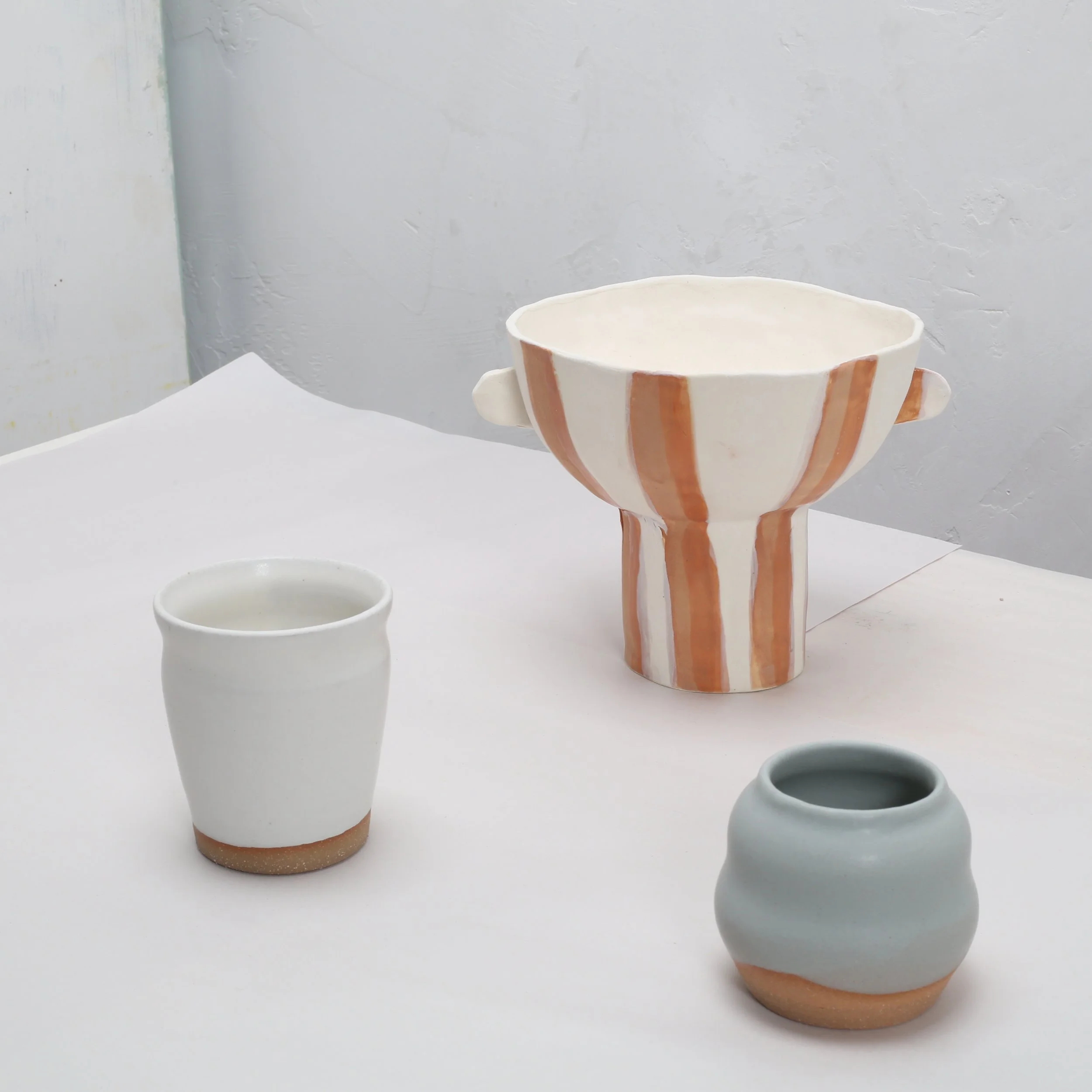 Three ceramic cups with a white glaze, each with orange-brown stripes. One cup is larger with a curved handle, one smaller without a handle, and the third is a medium-sized cup with a round opening. The cups are placed on a white surface against a light gray textured wall.