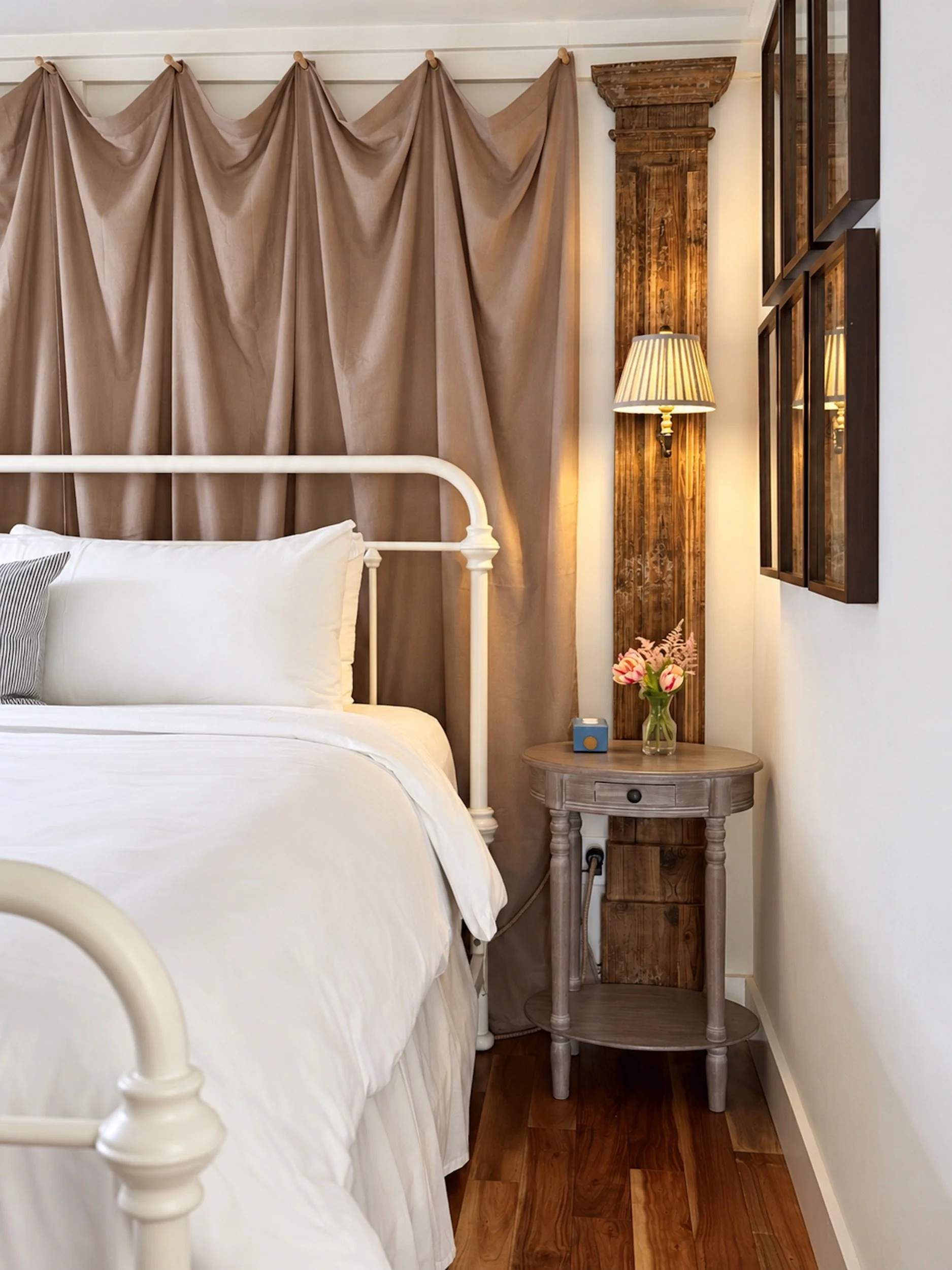 A detail of a bed with fabric hanging on the wal behind the bed, an antique column on the wall behind the bedside table and a wall sconce as bedside lamp