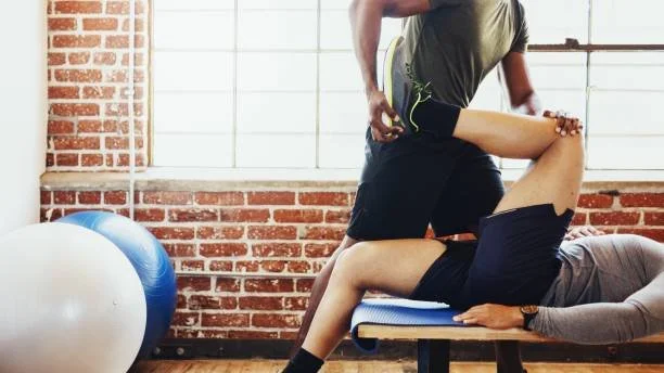 Person performing a stretching exercise on a bench while another person assists. A large exercise ball is visible in the background inside a gym.