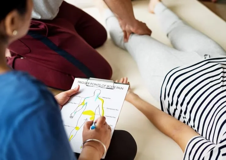 A healthcare professional taking notes on a diagram of body pain points during a medical consultation with a patient lying on an examination table.