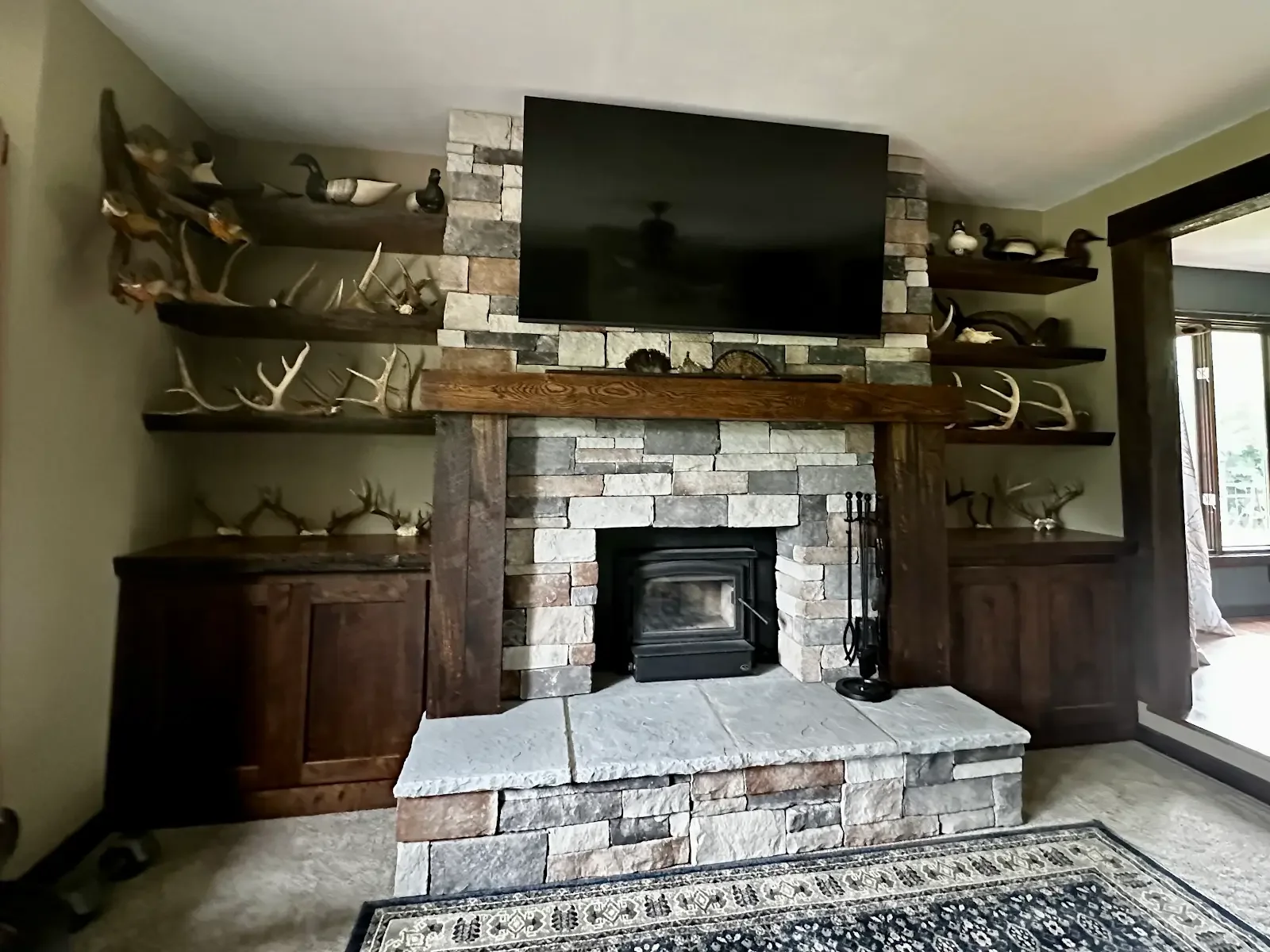 Living room fireplace with a stone surround, wooden mantel, and a mounted flat-screen TV above. Decor includes mounted antlers on dark wood shelves on either side of the fireplace.