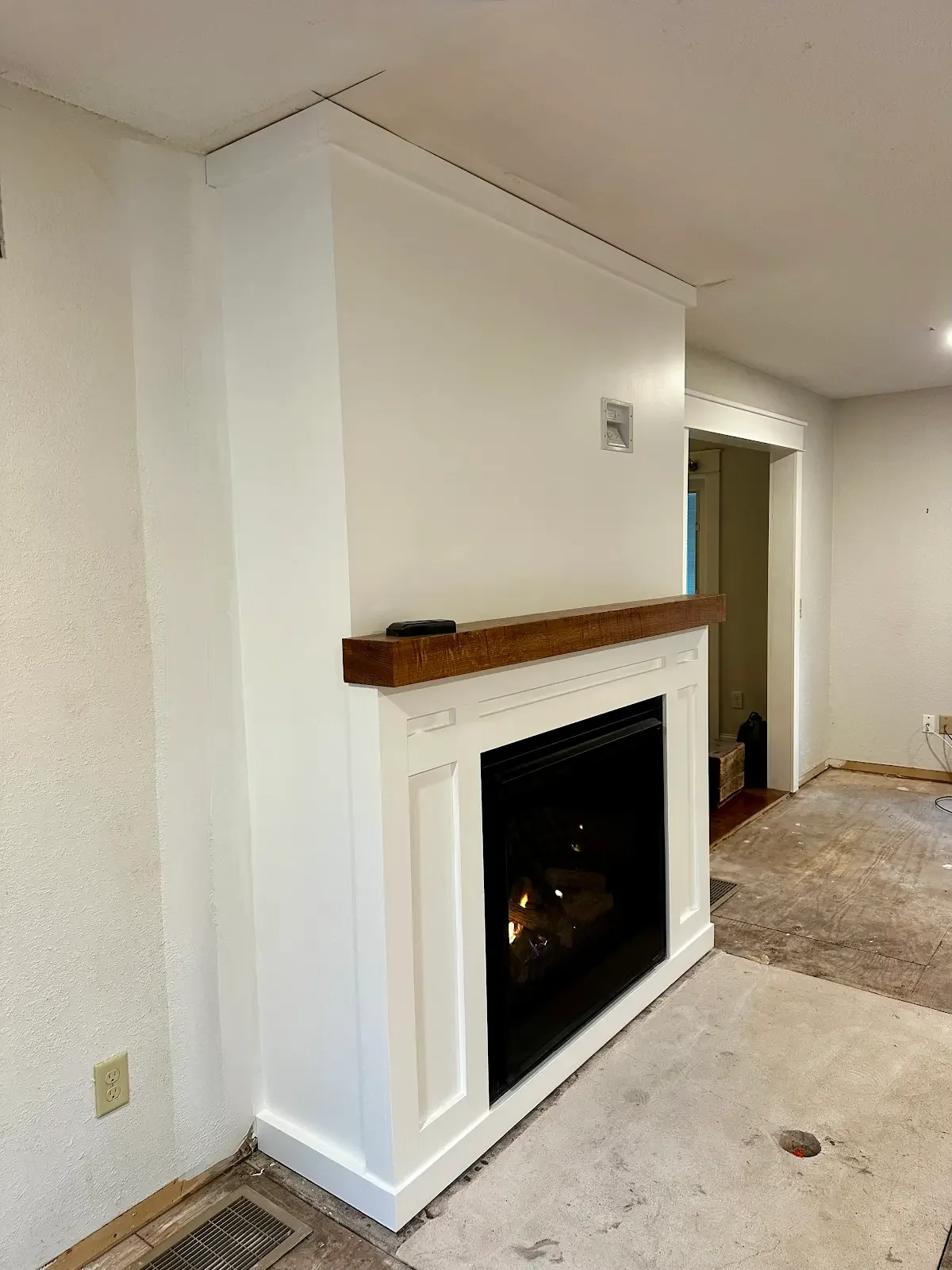 Living room with a white wall and fireplace, a wooden mantel, and a small object on the mantel. The floor appears to be under renovation, with exposed subfloor and an electrical outlet on the wall.
