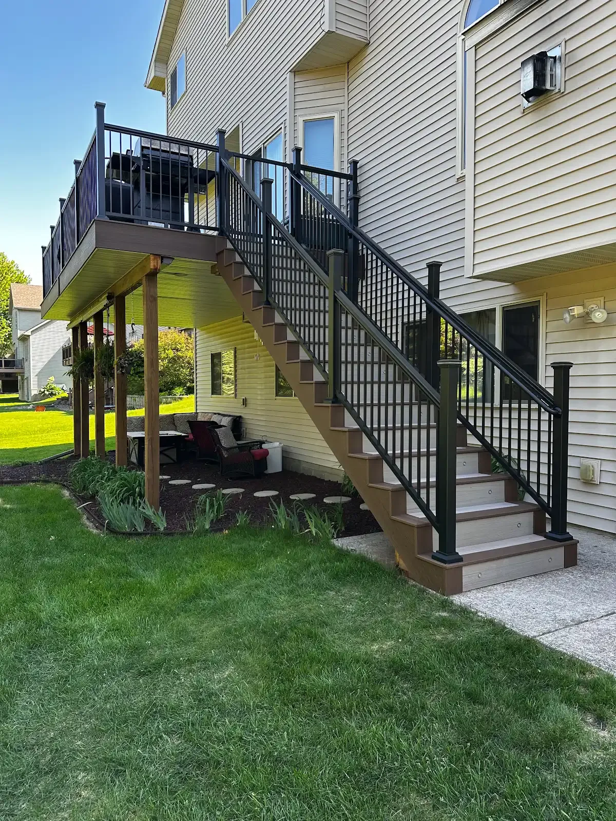 Exterior view of a multi-story house with a staircase leading to a second-floor balcony, a backyard patio with seating, green lawn, and neighboring houses in the background.