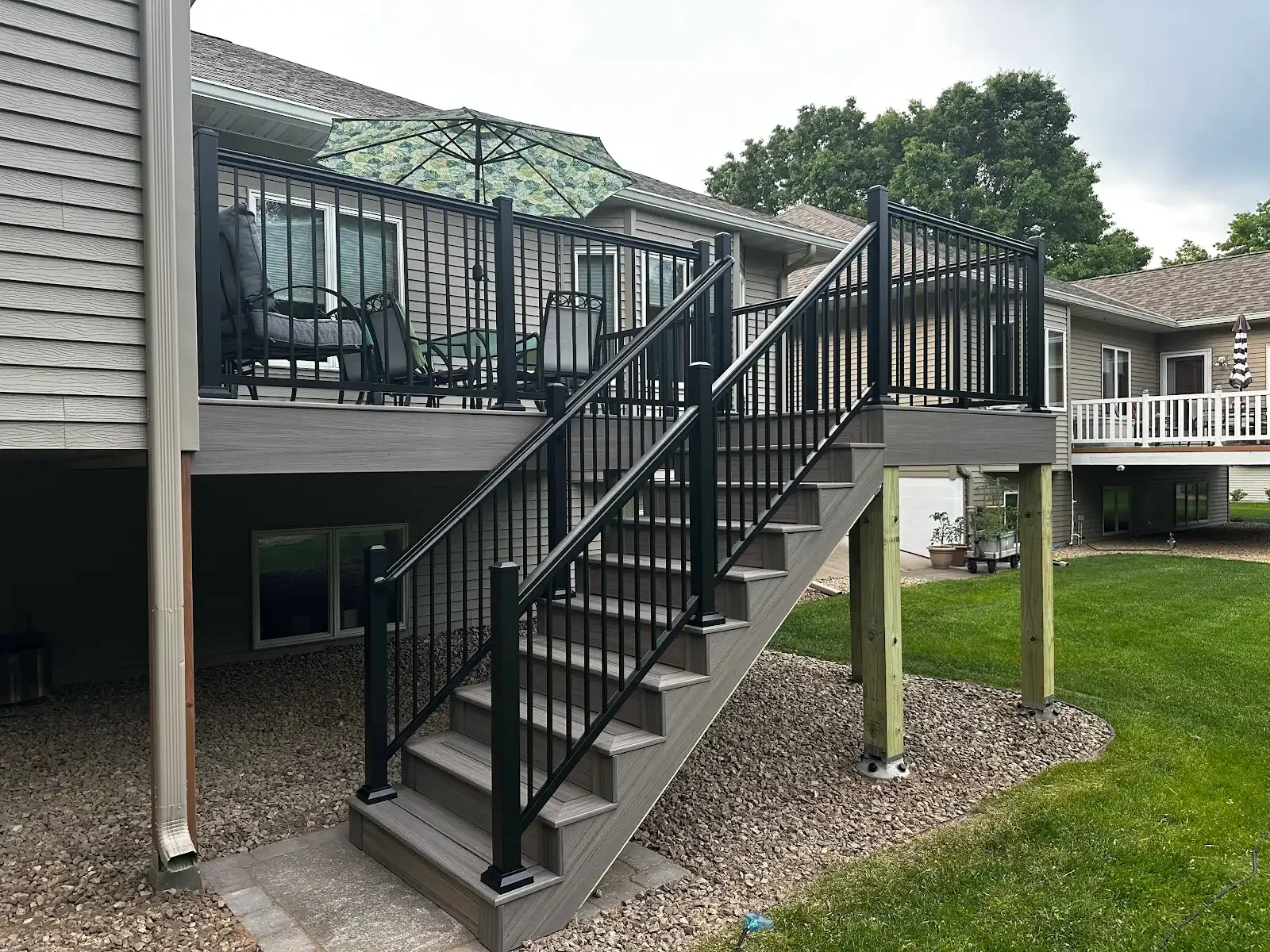 A backyard deck with stairs leading down to a grassy lawn, featuring a black metal railing, a patio table with chairs, and a glass patio umbrella.
